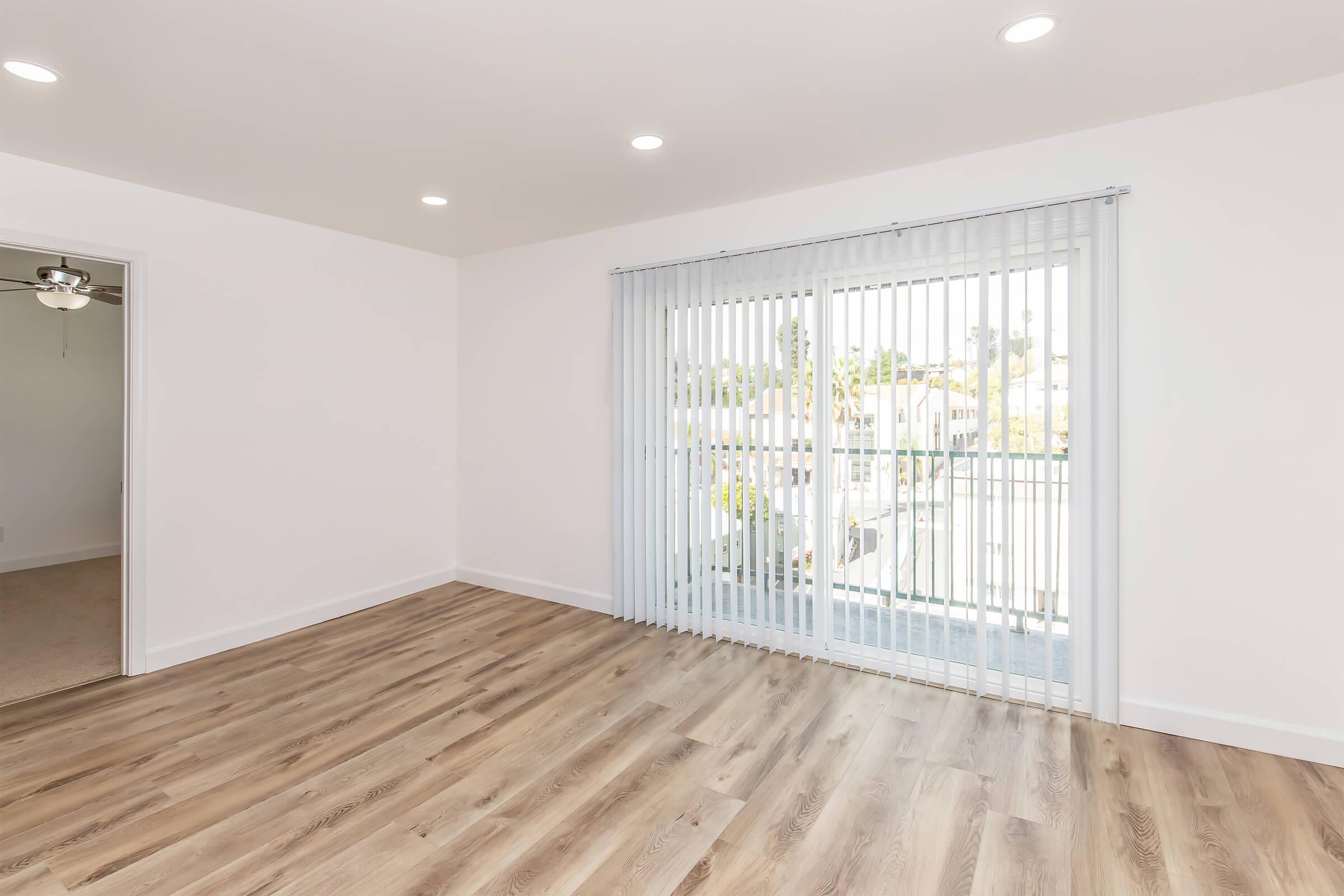 A bright, spacious interior featuring light wood flooring, white walls, and recessed lighting. Large sliding glass doors with vertical blinds lead to an outdoor area, allowing natural light to fill the room. A doorway on the left suggests access to another space.