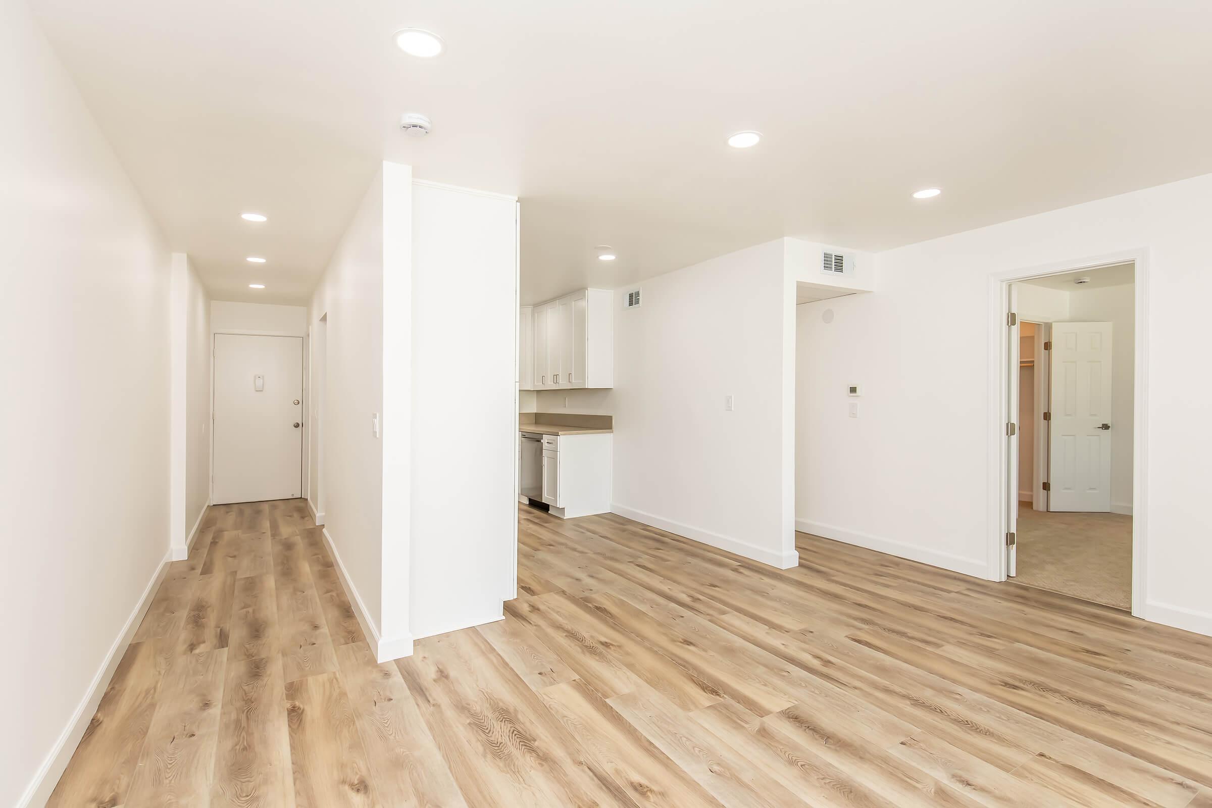 A modern, empty hallway with light-colored walls and hardwood flooring. The space features recessed lighting and leads to an open kitchen area with white cabinetry. A door on the right opens to a room, while the main entrance is visible at the far end of the hallway. The overall atmosphere is bright and spacious.