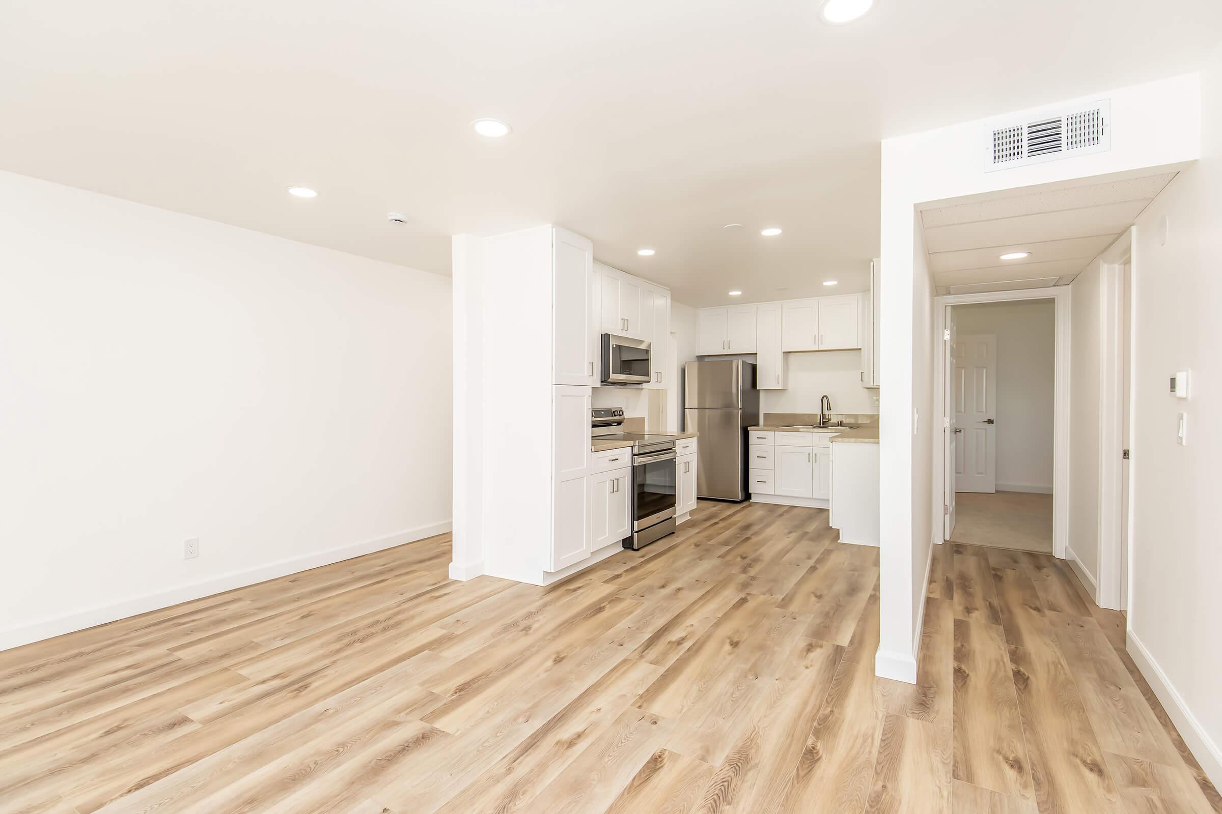 A spacious, modern kitchen and living area featuring wood-style flooring, white cabinets, stainless steel appliances, and ample natural light. The layout includes an open design connecting the kitchen to the living space, with clean lines and a neutral color palette.