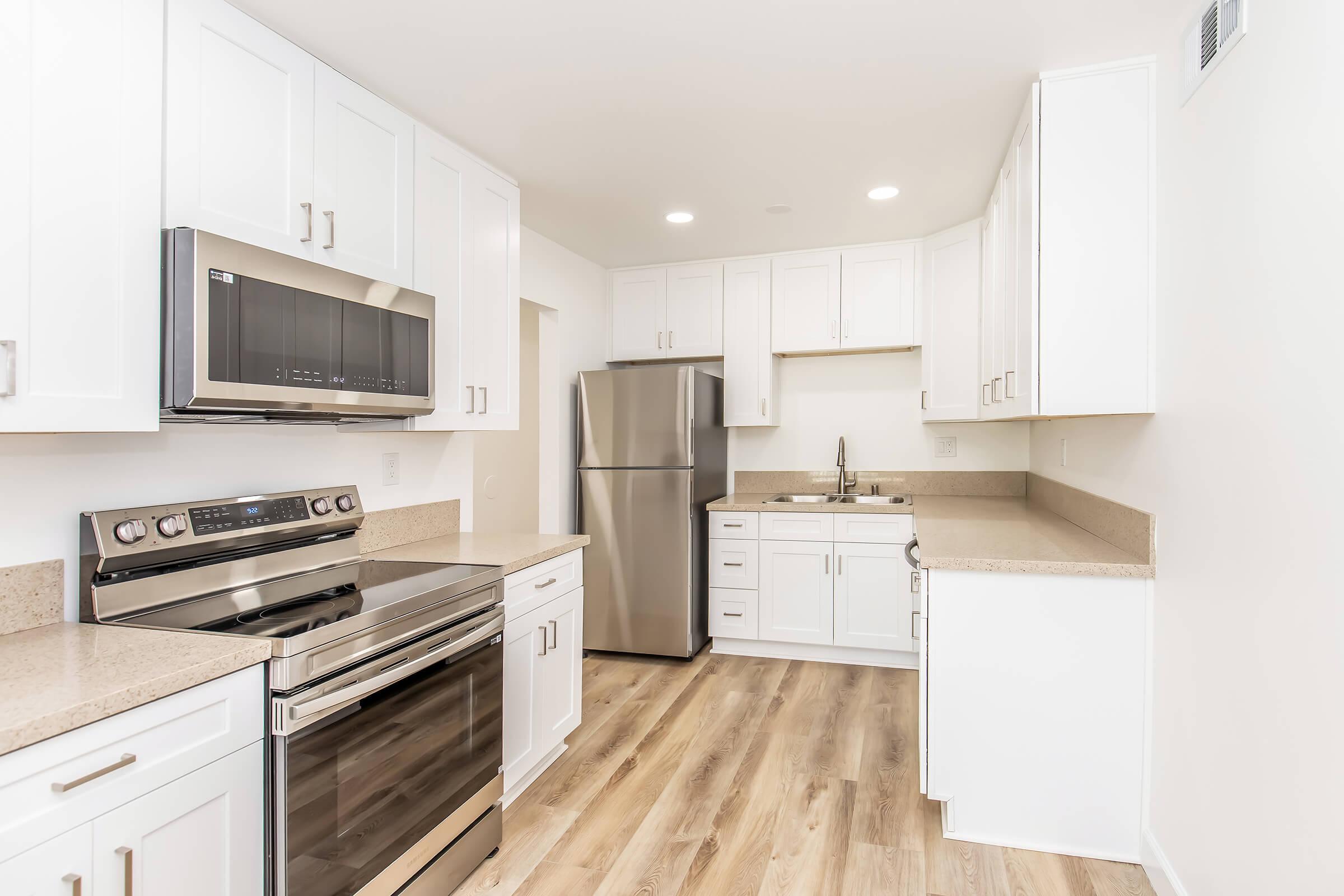 Modern kitchen featuring white cabinets, stainless steel appliances including an oven, microwave, and refrigerator, and a light-colored countertop. The flooring is made of light wood, and the space is well-lit with recessed lighting. The overall design is clean and contemporary.