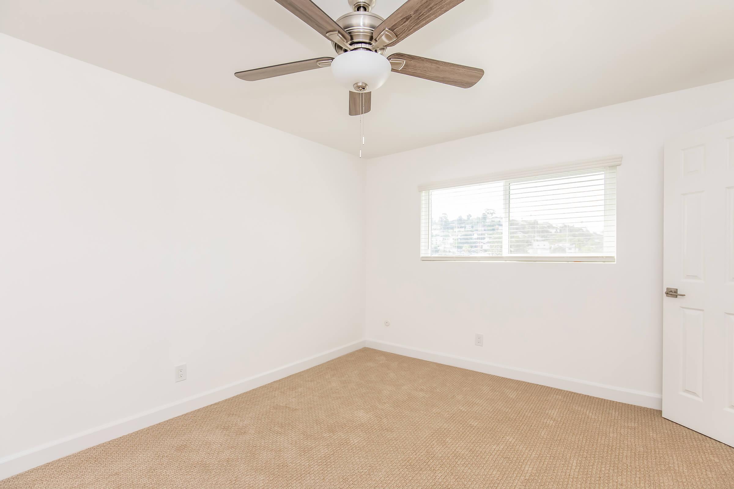 A bright, unfurnished room with light beige carpet and white walls. There is a ceiling fan with wooden blades and a central light fixture. A window with white blinds lets in natural light, while a closed door is visible on the right. The room has a minimalist and clean appearance, suitable for various uses.