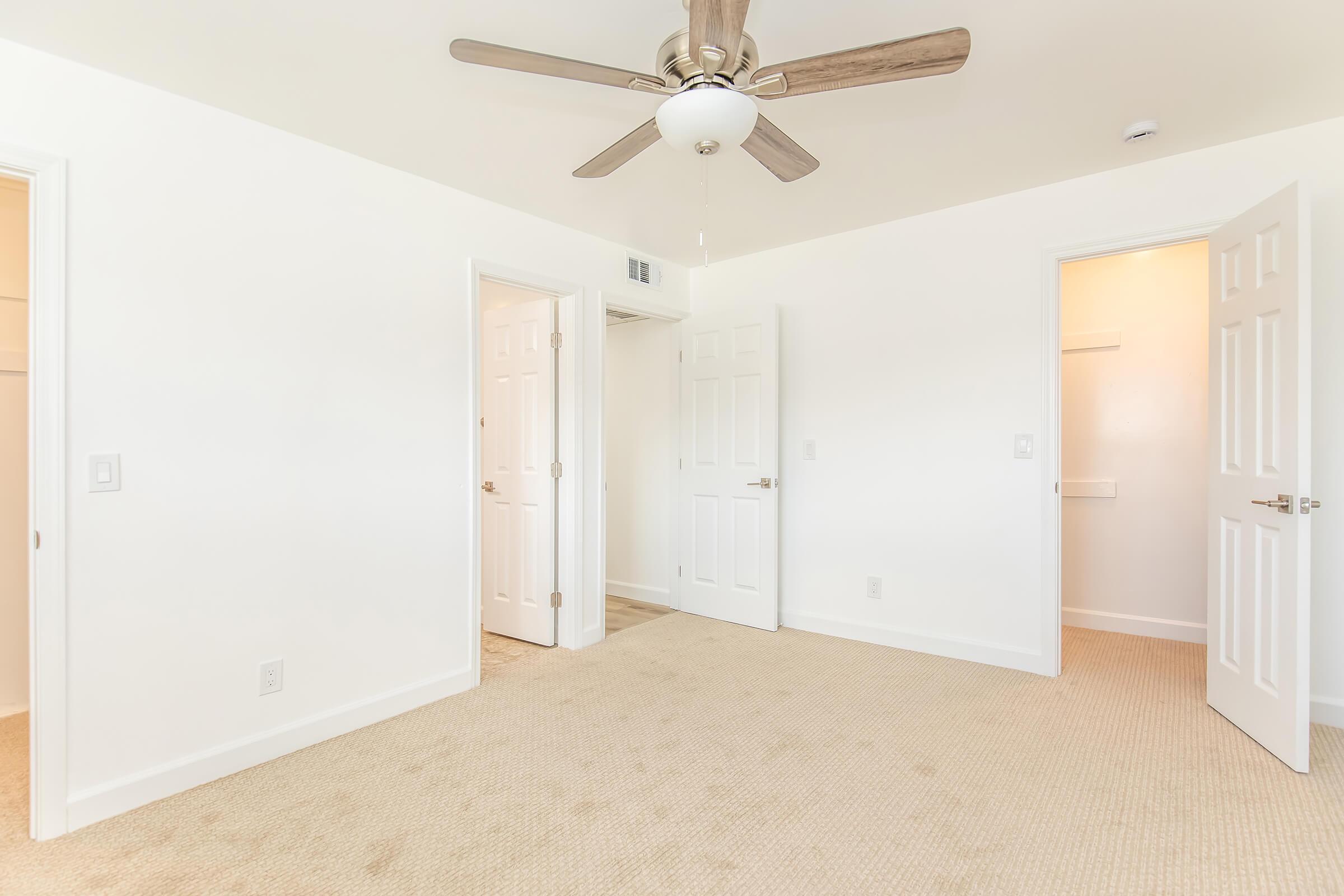 A bright and empty room featuring beige carpet, white walls, and a ceiling fan. There are two closed doors on opposite walls, leading to adjacent spaces, and one open door revealing a small closet. The room is well-lit, creating an airy atmosphere.