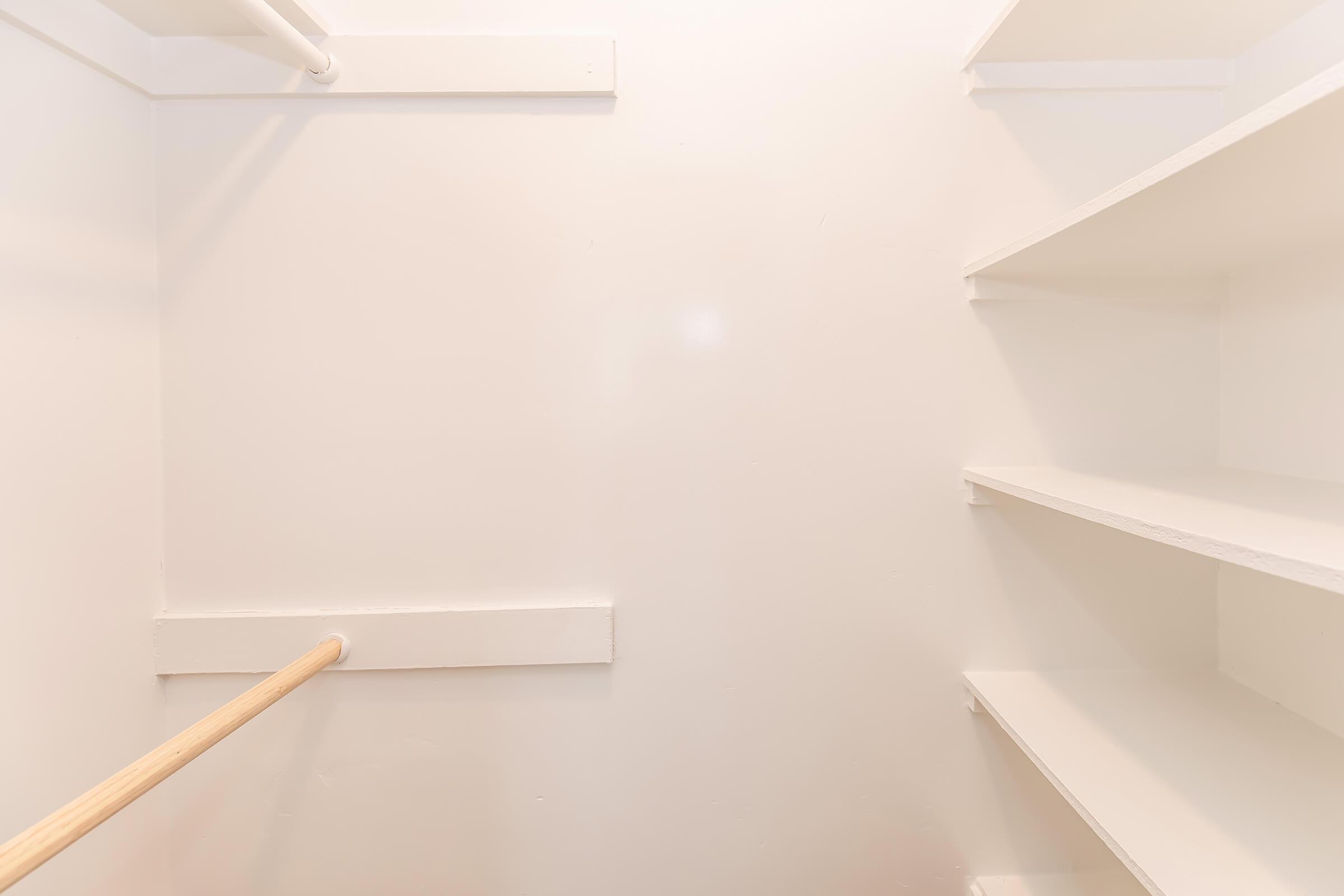 A clean, empty closet featuring a wooden hanging rod on the left side and white shelves on the right against a white wall. The space is well-lit and organized, with no items or clutter visible.