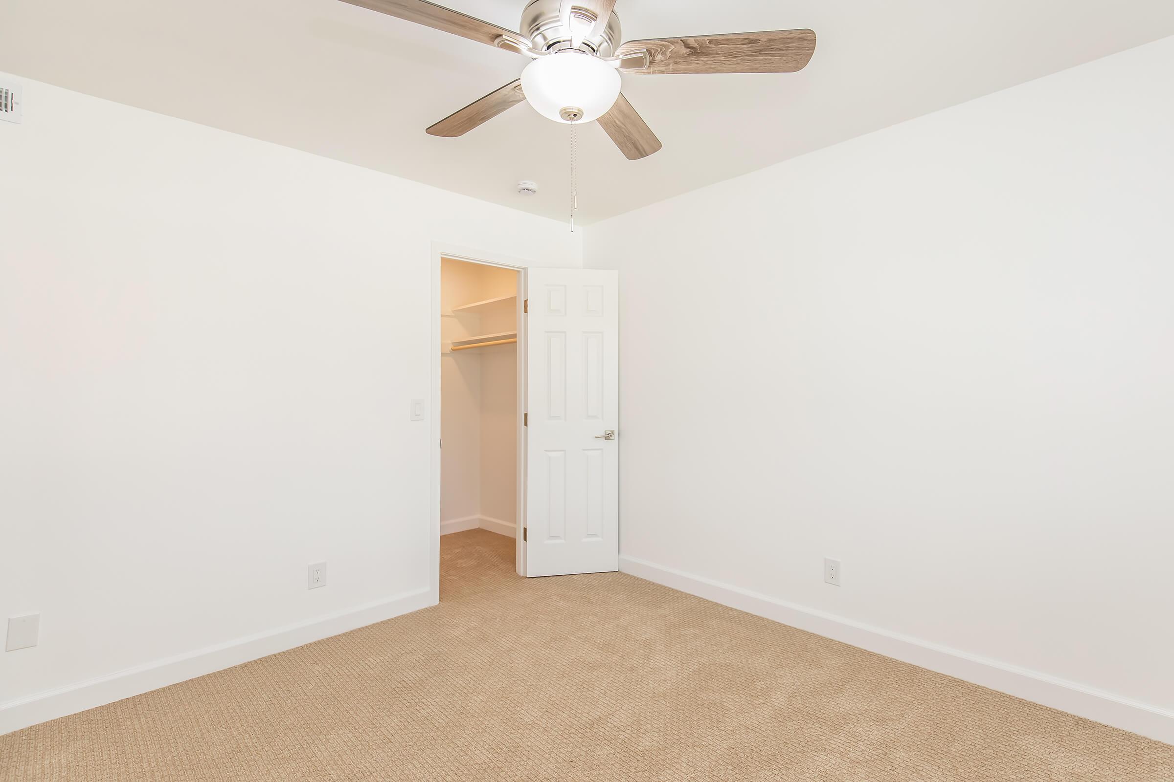 Bright and empty room featuring a ceiling fan and a light fixture. The walls are painted white, with a beige carpeted floor. An open door leads to a small closet area.