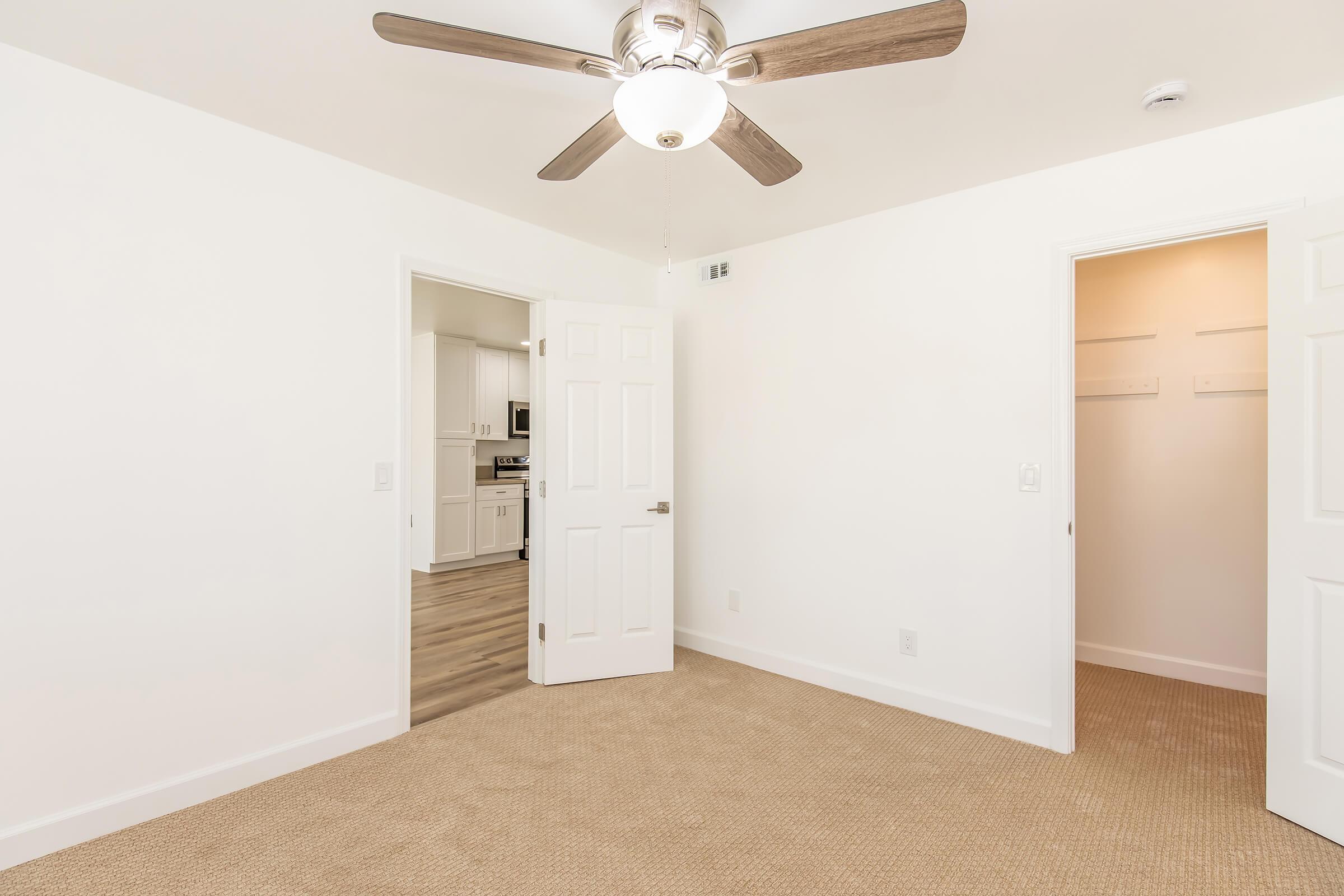 A well-lit room with a ceiling fan, featuring two white doors leading to adjacent areas. The floor is carpeted in a neutral color, and the walls are painted white, creating a clean and modern appearance.