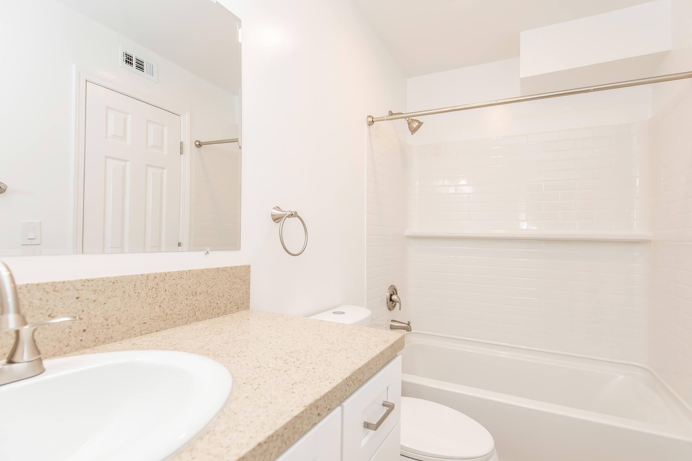 A clean and modern bathroom featuring a white bathtub with a shower, a beige countertop with a sink, a round mirror above the sink, and a towel ring on the wall. The walls are painted white, and a door is visible in the background. The space appears bright and well-lit.