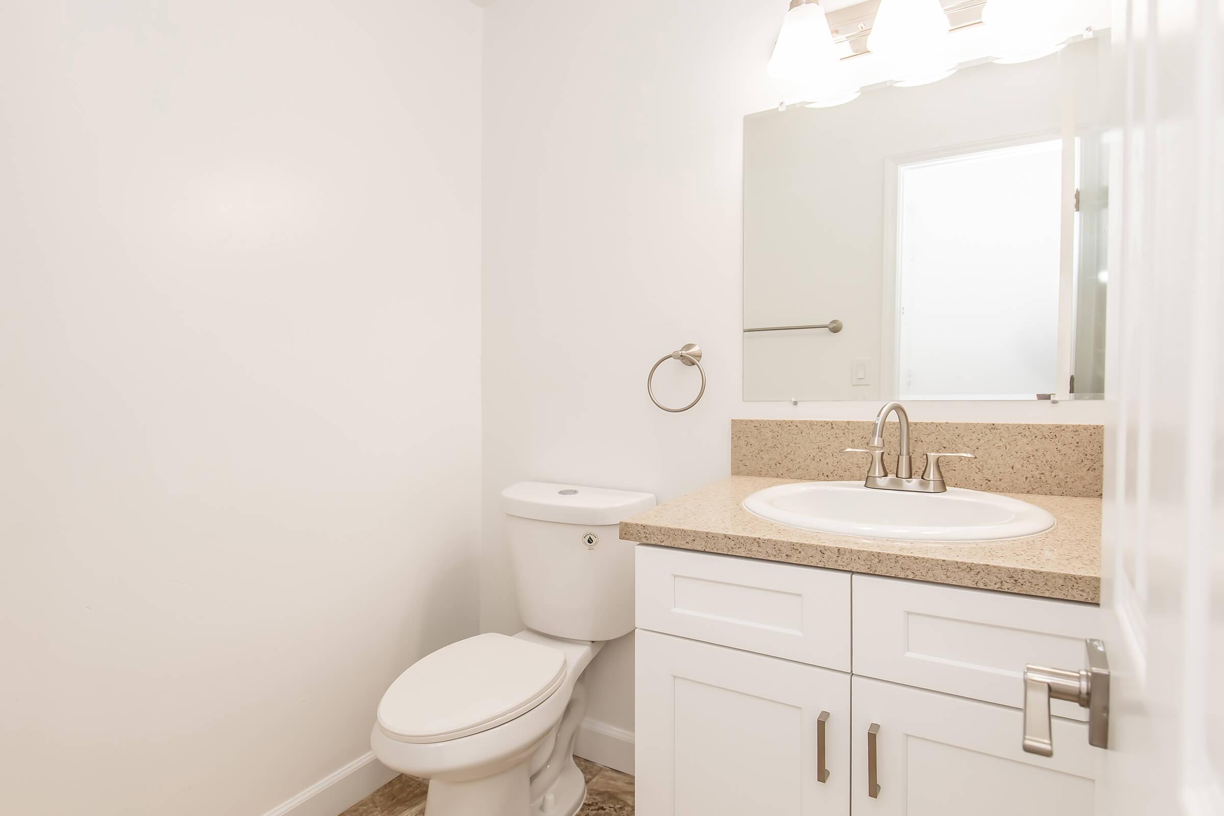 A clean, modern bathroom featuring a white toilet, a sink with a granite countertop, and a mirror above it. The walls are painted white, and there is a towel bar next to the sink. Natural light enters from a nearby door. The overall design is simple and contemporary.
