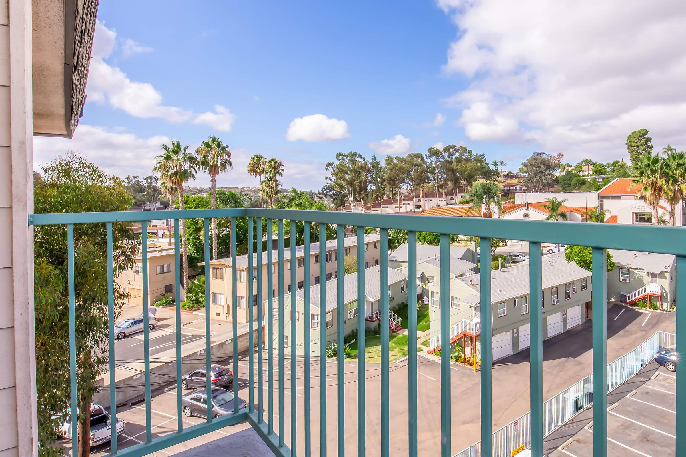 A view from a balcony overlooking a residential area, featuring green palm trees, multiple buildings with varying architectural styles, and a clear blue sky with scattered clouds. The scene captures a sunny day, showcasing a tranquil neighborhood atmosphere.