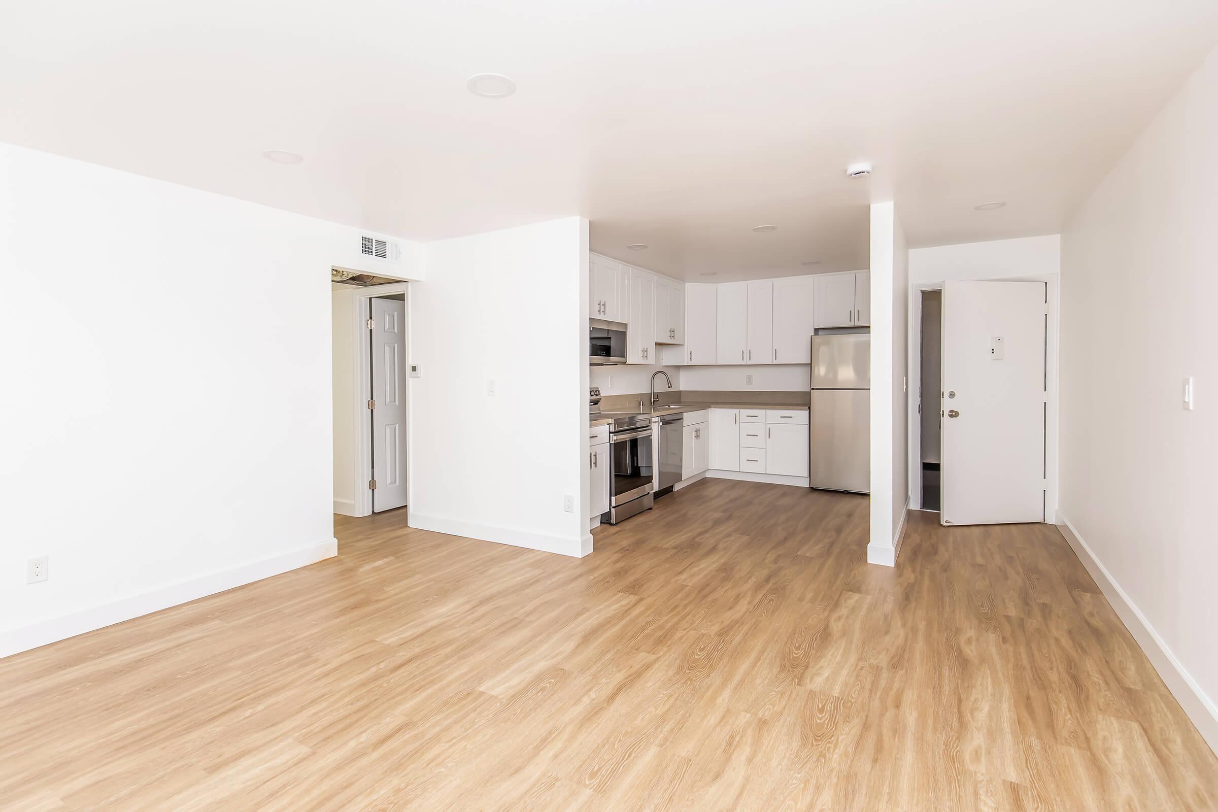 A modern, empty kitchen and living space with light wood flooring, white walls, and stainless steel appliances. The kitchen features an open layout with cabinets and a countertop. A door leads to another room, and there's an entrance door visible on the right. Brightly lit with natural light.