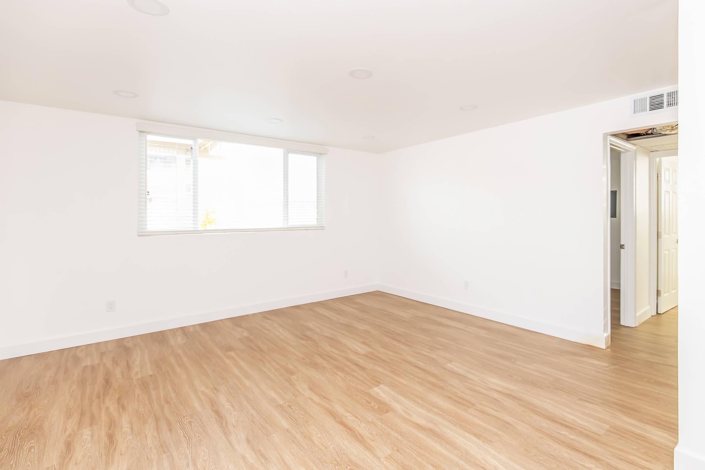 A spacious, empty room with white walls and light wood flooring. A large window allows natural light to enter, illuminating the space. There is a doorway on the right leading to another area, and the ceiling features recessed lighting. The overall ambiance is bright and minimalistic.
