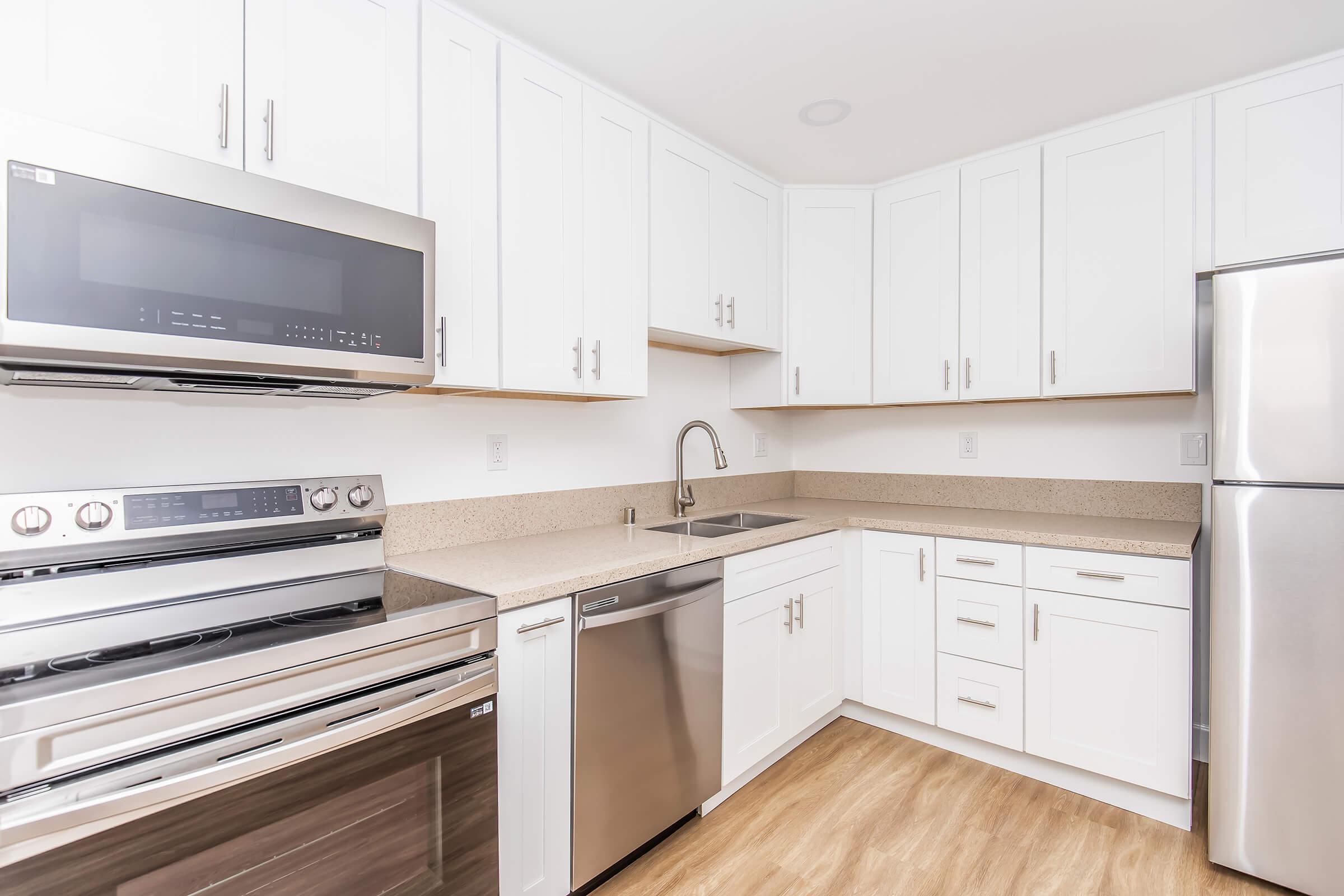 Modern kitchen featuring white cabinets, stainless steel appliances including a microwave and oven, a sleek sink with a faucet, and a dishwasher. The countertop is beige with a subtle pattern, and the flooring is a light wood finish. The kitchen is well-lit with a clean, contemporary design.