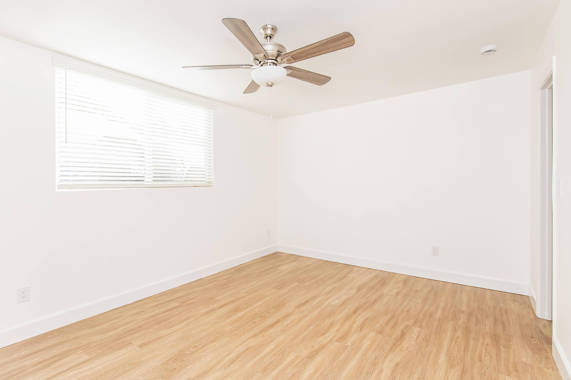 A bright, empty room with white walls and wooden flooring. A ceiling fan is centrally located, and there is a window with blinds allowing natural light to enter. The space has a minimalist design, suitable for various uses, with a door visible on the right side.
