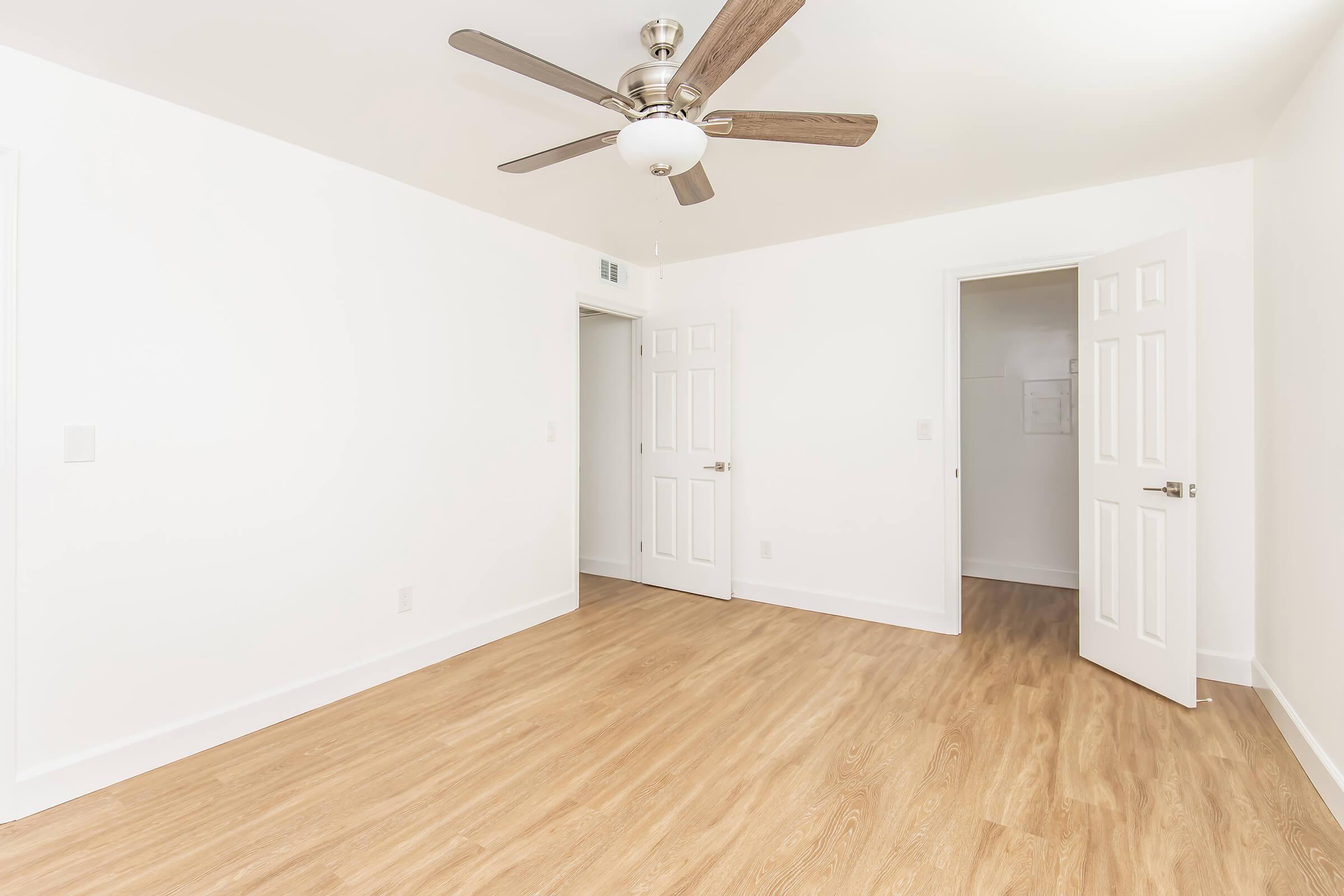 A bright, empty room with light-colored walls and a light wood floor. A ceiling fan is visible in the center, and two white doors lead to adjacent spaces. The overall ambiance is clean and airy, creating an impression of spaciousness.