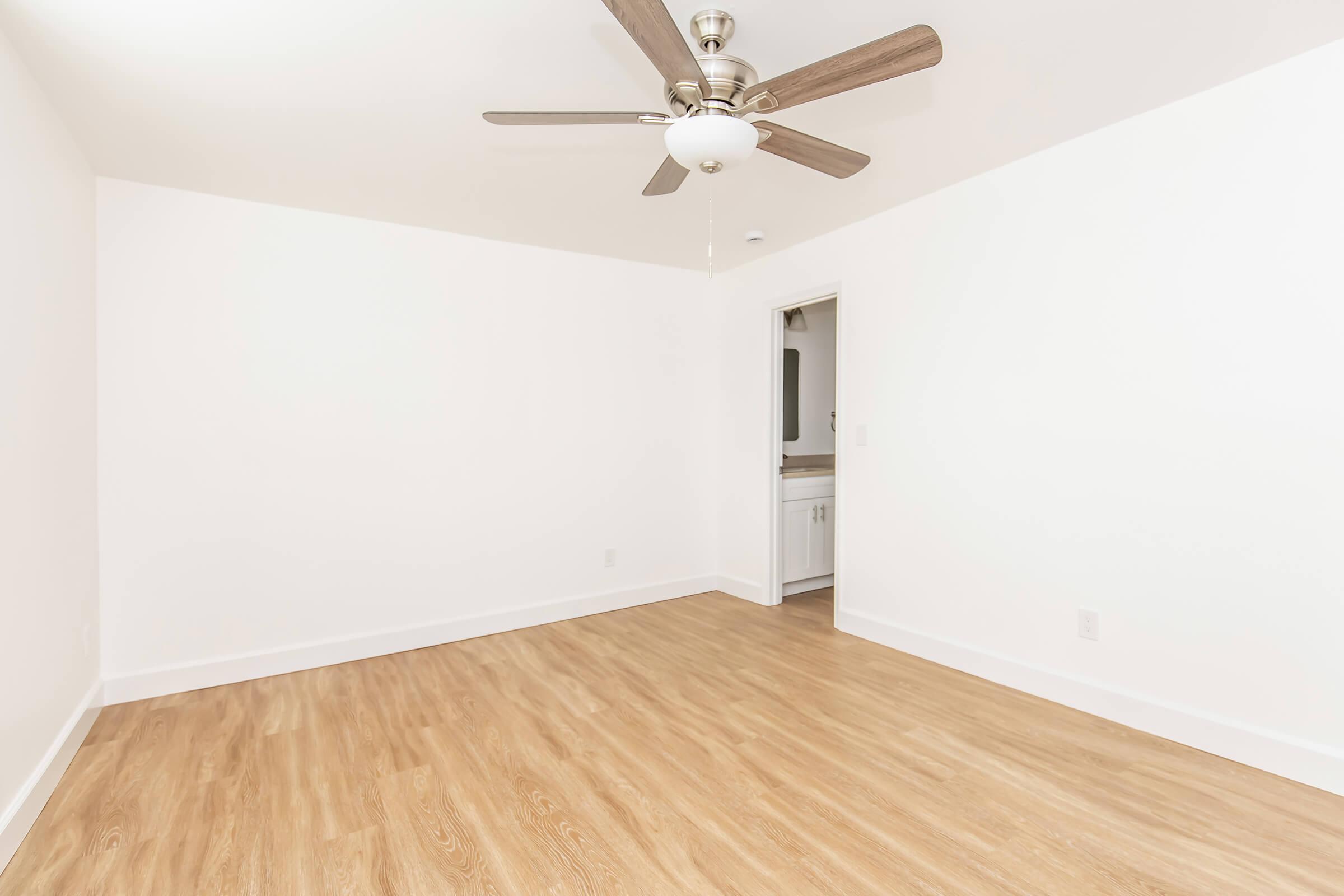 Empty room with light wood flooring, a ceiling fan with wooden blades, and white walls. A doorway can be seen on the right, leading to another space. Natural light enhances the bright, minimalist feel of the room.