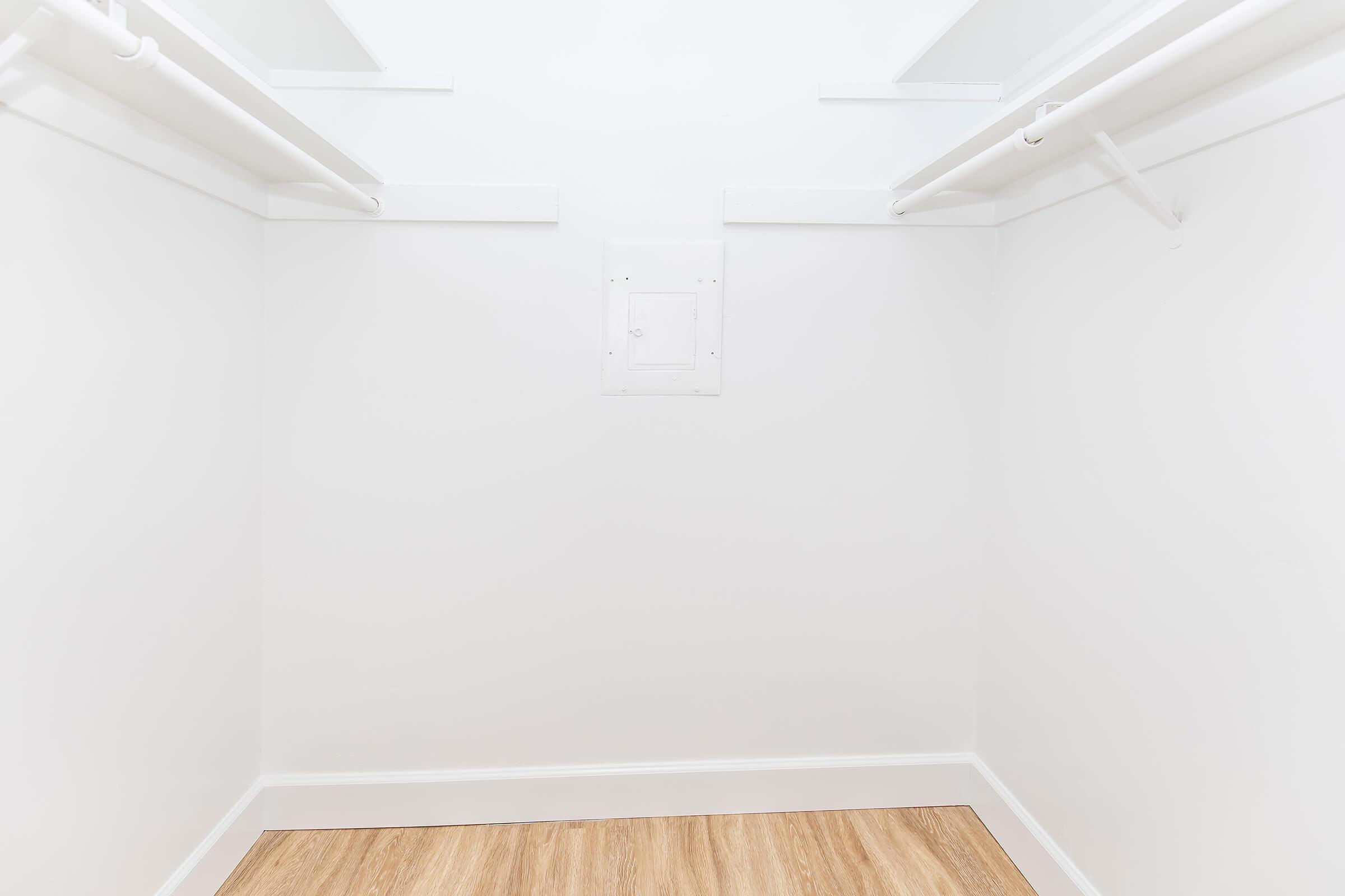 A spacious, empty walk-in closet featuring white walls, two hanging rods on either side, and a wooden floor. The closet has a small access panel on the back wall, providing a minimalist and clean aesthetic. Ideal for storage or organization purposes.