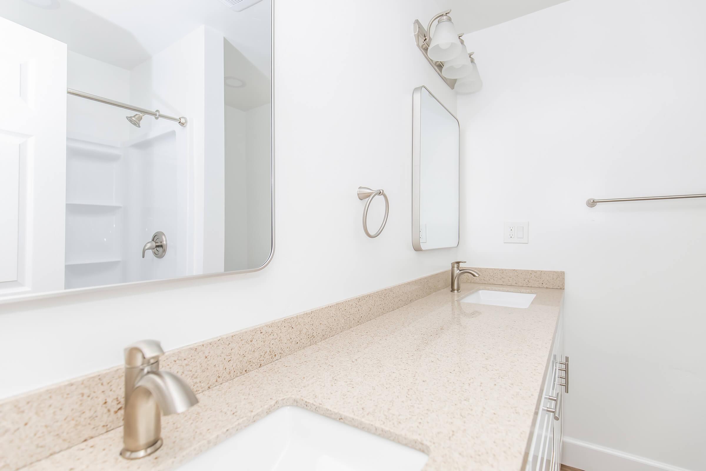 A clean and modern bathroom featuring a double sink vanity with a beige countertop, a large wall-mounted mirror, and a towel ring. The shower area is positioned behind a frosted glass panel, and the walls are painted white, contributing to a bright, airy atmosphere.