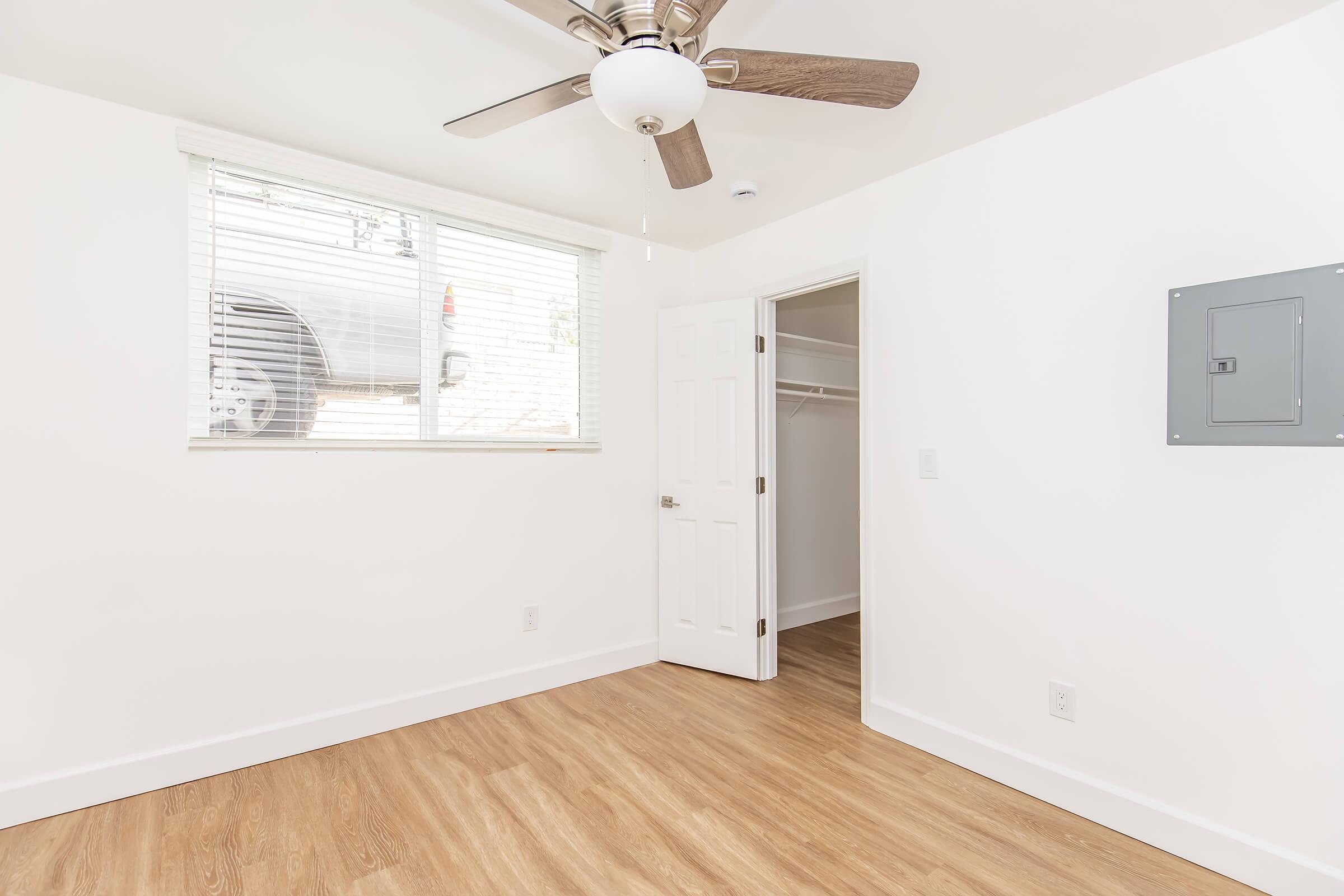 Empty room featuring a ceiling fan, light-colored walls, and a large window with blinds. A door leads to a closet, and there is an electrical panel mounted on the wall. The flooring is light wood, creating a modern and clean aesthetic.
