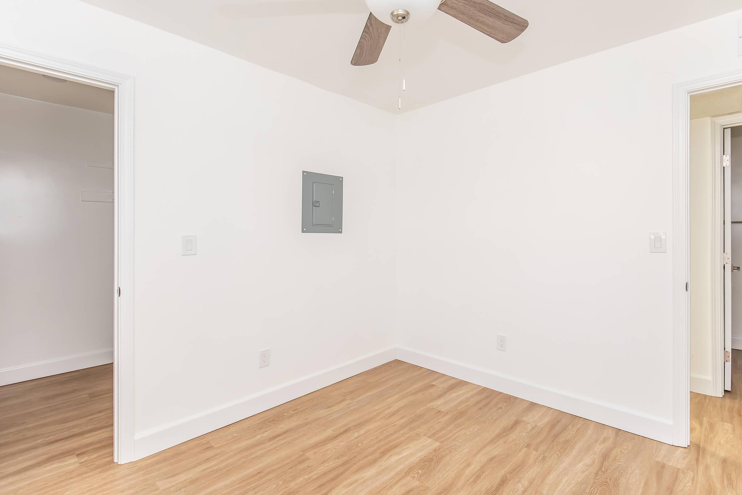 Empty room with white walls and a ceiling fan. There are two doorways on opposite walls, and an electrical panel is mounted on one wall. The floor is wooden, creating a bright and airy atmosphere. Overall, the space appears clean and minimalist.