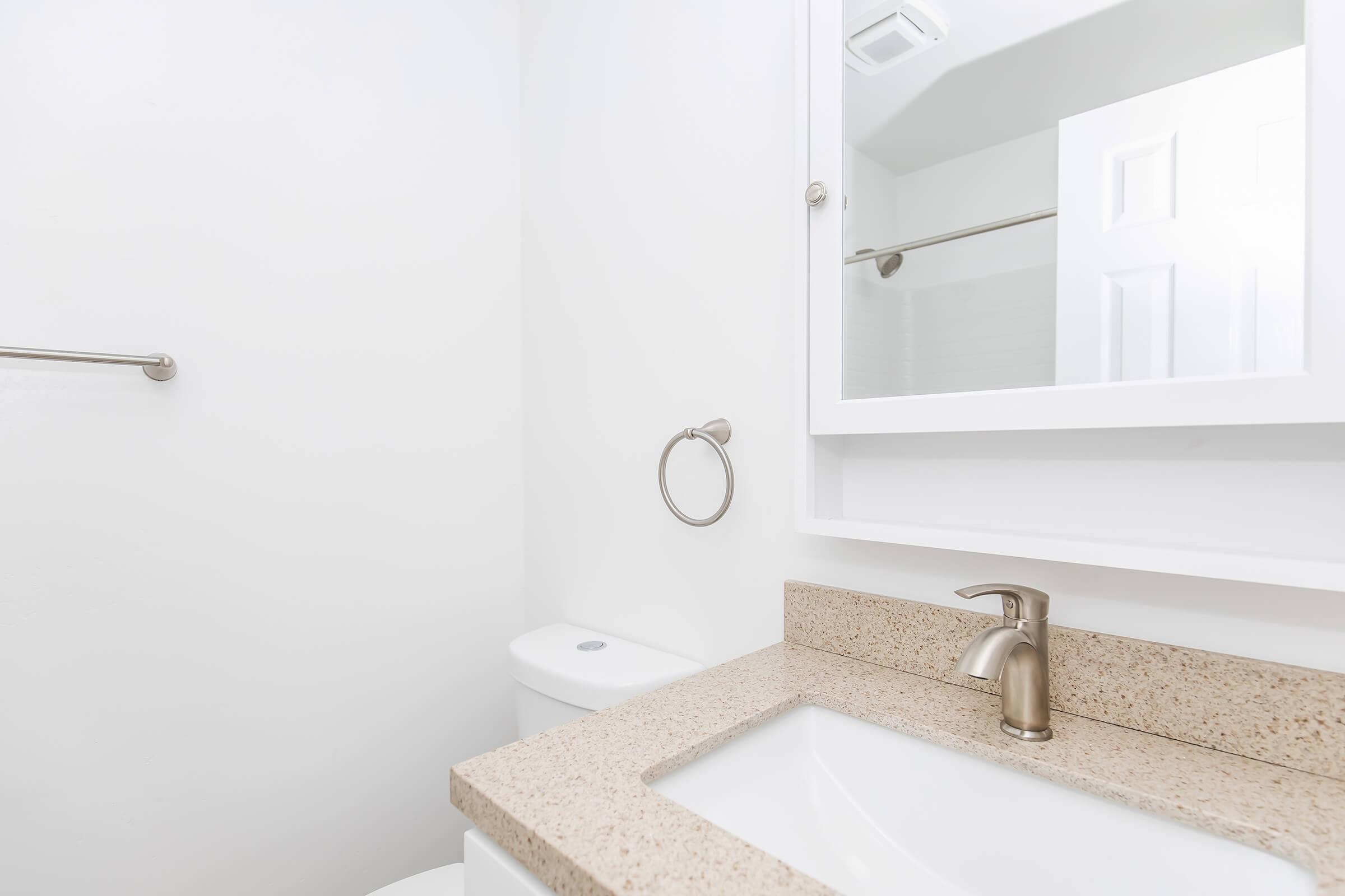 A bright, modern bathroom featuring a white sink with a sleek faucet, beige granite countertop, and a wall-mounted mirror. A toilet is visible in the background, along with a towel bar and a hand towel ring. The overall color scheme is clean and minimalistic, with white walls and neutral tones.