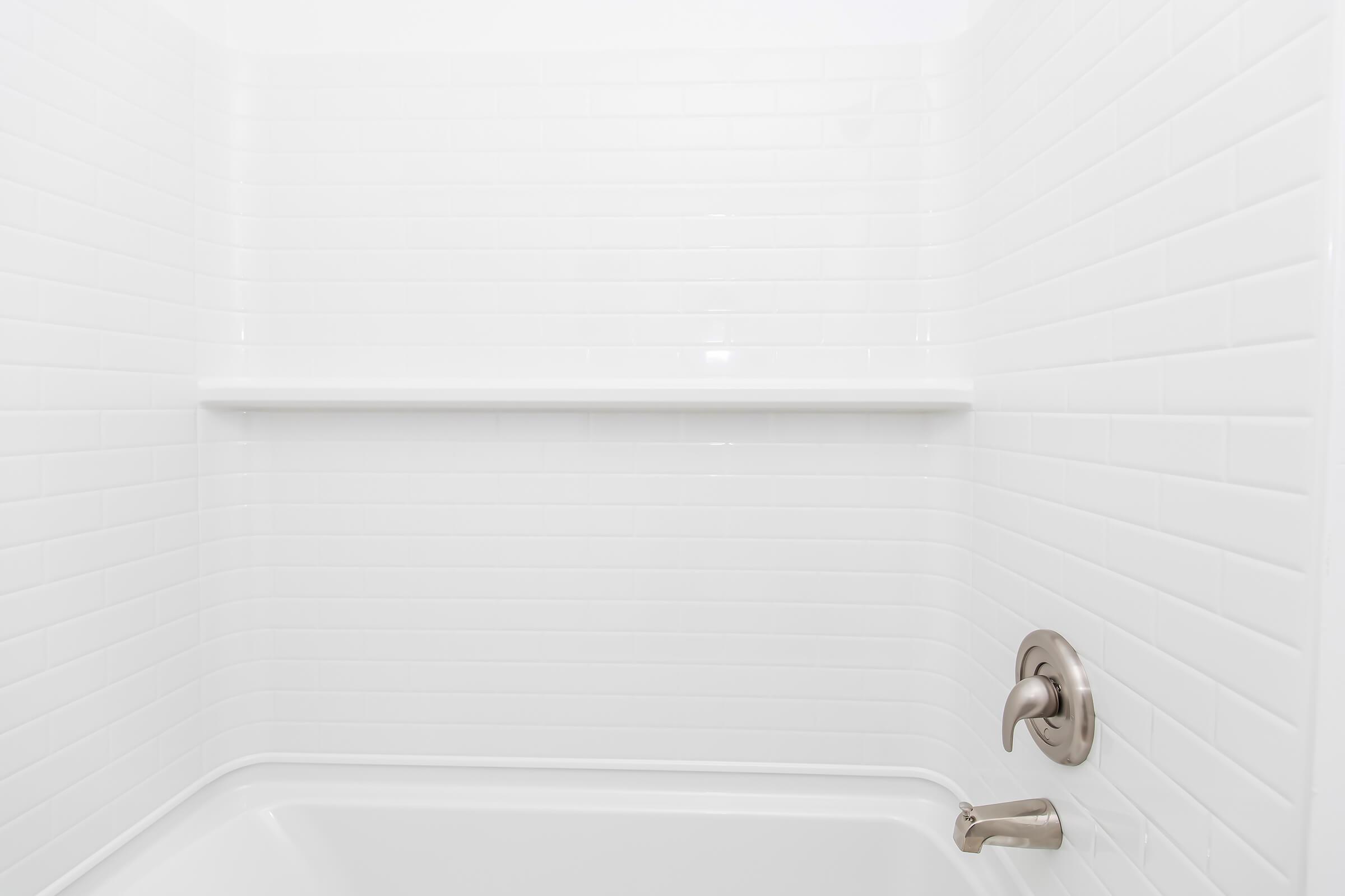 Clean, minimalistic bathroom shower area with white tiled walls, featuring a simple shelf and a modern faucet and control handle. The overall design is bright and uncluttered, emphasizing a fresh and serene atmosphere.