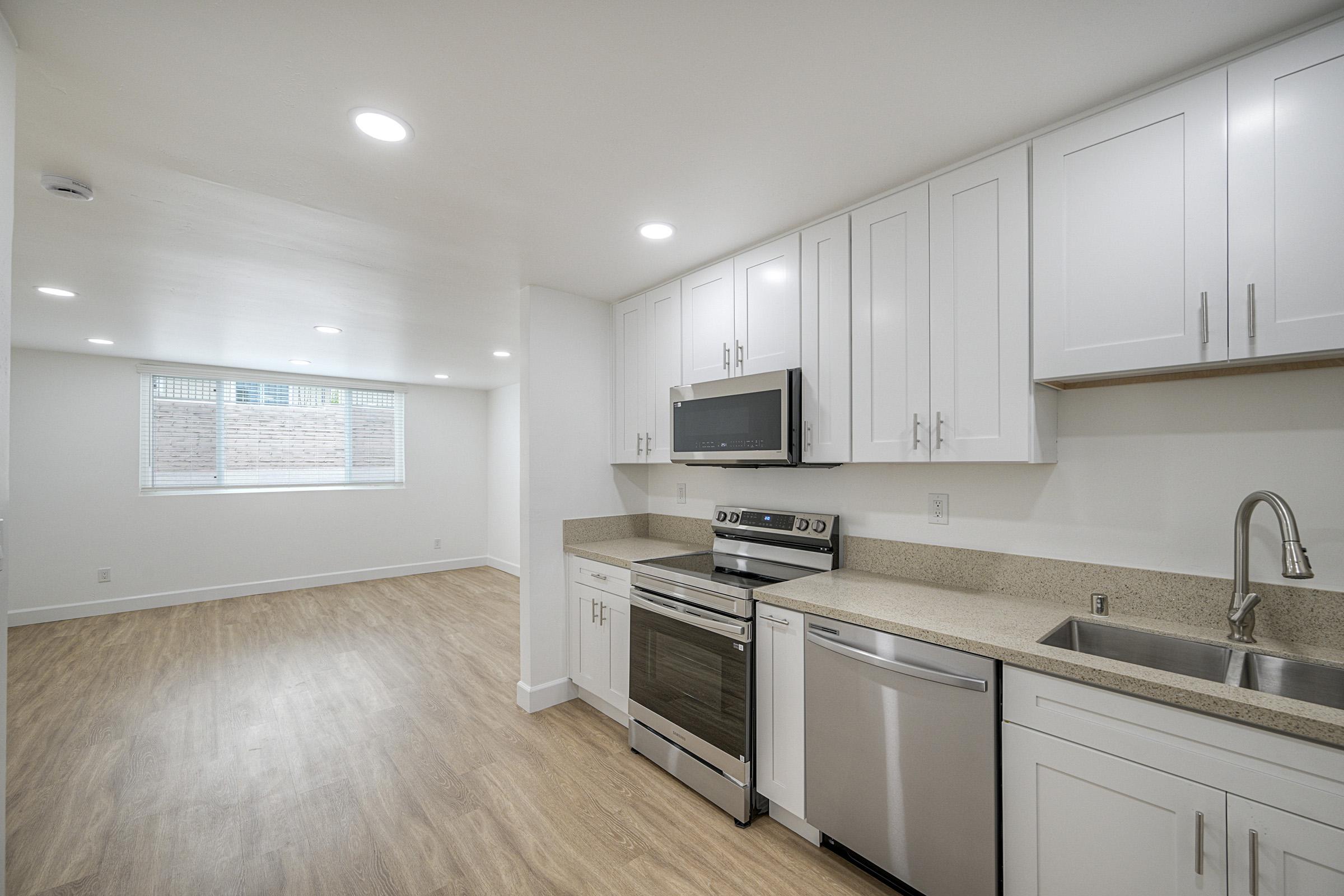 Bright, modern kitchen with white cabinets and stainless steel appliances, including an oven and microwave. The space features a neutral color palette, sleek countertops, and a window allowing natural light. Hardwood-style flooring extends into the adjacent living area.