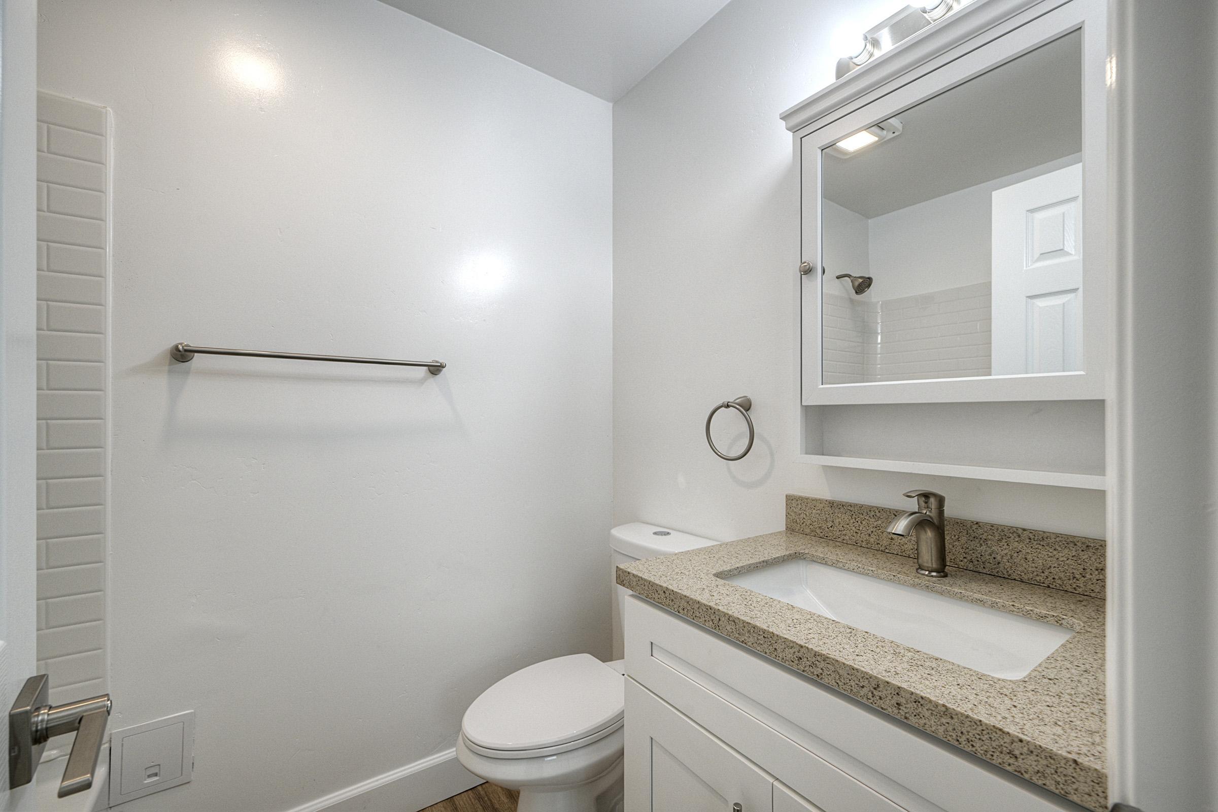 A modern bathroom featuring a white wall, a cabinet with a sink, a large mirror, a toilet, and a towel rack. The countertop is made of a light-colored stone, and the flooring appears to be wood. The space is well-lit, creating a clean and bright atmosphere.