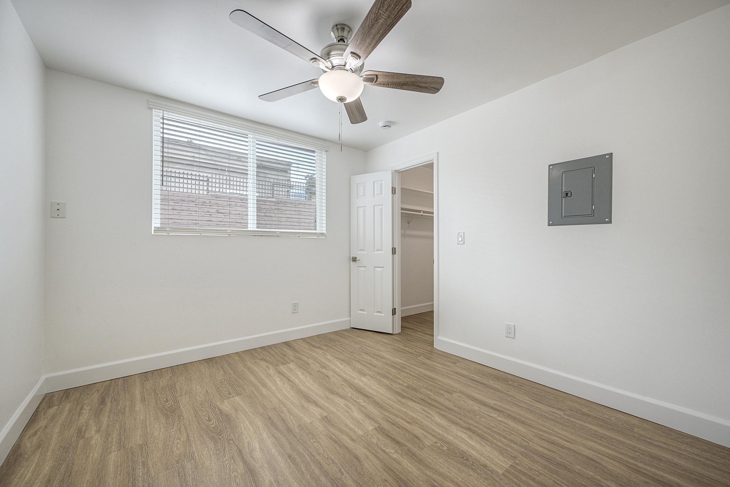 Empty room with light-colored walls and a ceiling fan. A window lets in natural light, and there's a door leading to a closet. The floor is wooden laminate, and an electrical panel is mounted on the wall. The overall space is bright and uncluttered.