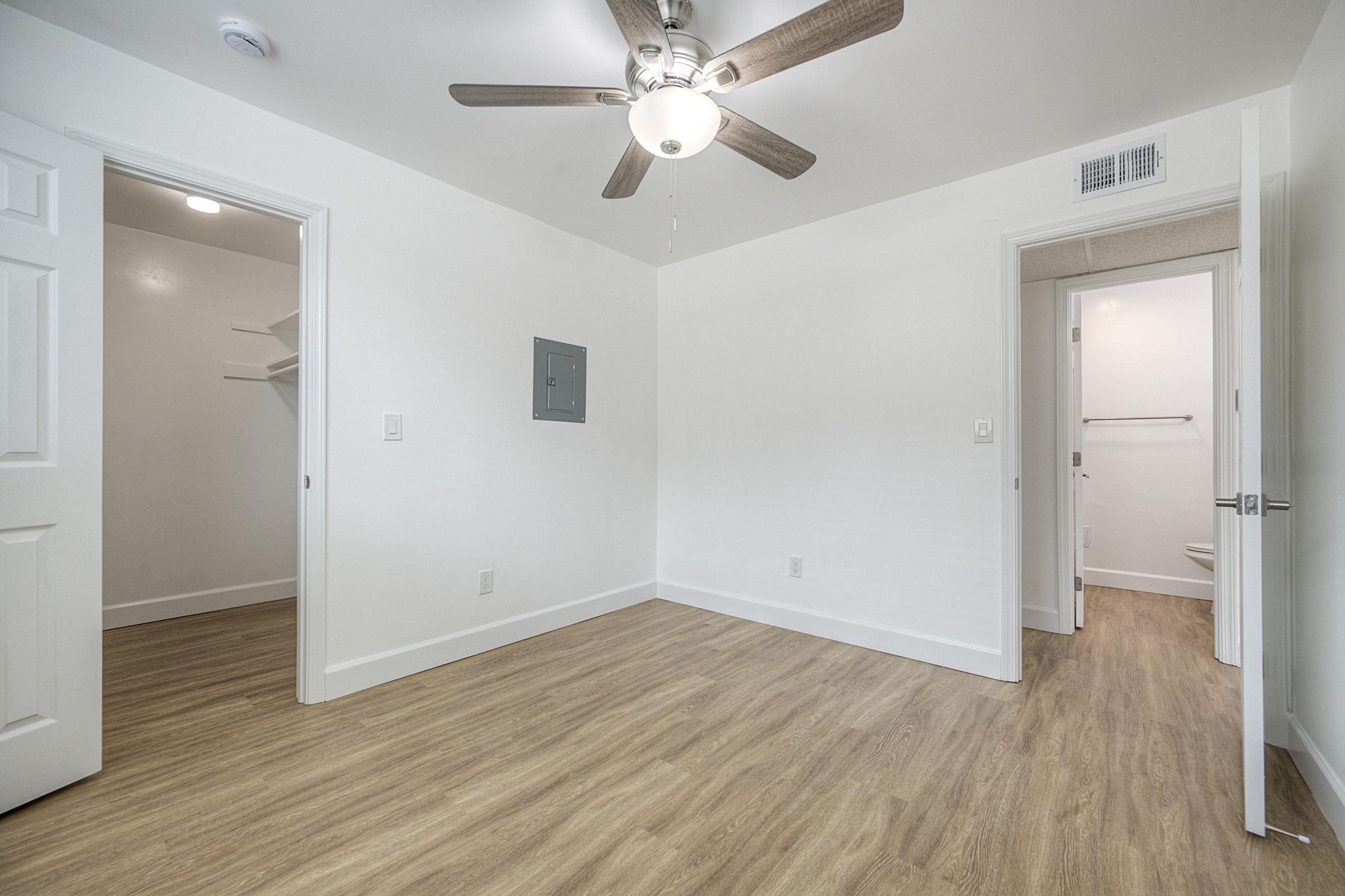A spacious room with light-colored walls and wooden flooring. It features a ceiling fan, a closet on the left, a wall-mounted electrical panel, and a door leading to a bathroom. The area is well-lit with natural light, providing a clean and modern aesthetic.
