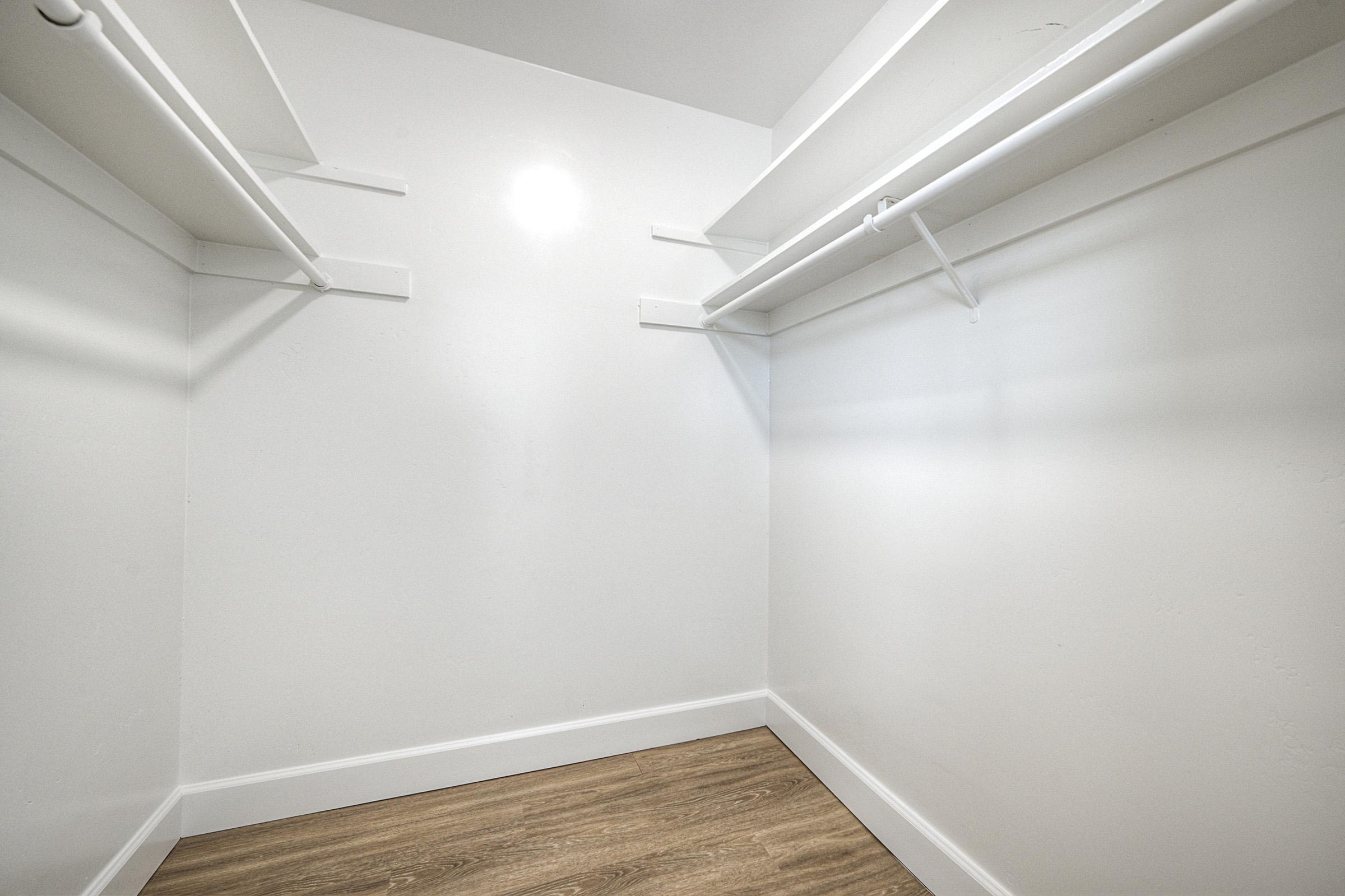 Empty walk-in closet featuring white walls, two hanging rods on either side, and a wooden floor. The space is well-lit with no additional furniture or items present.
