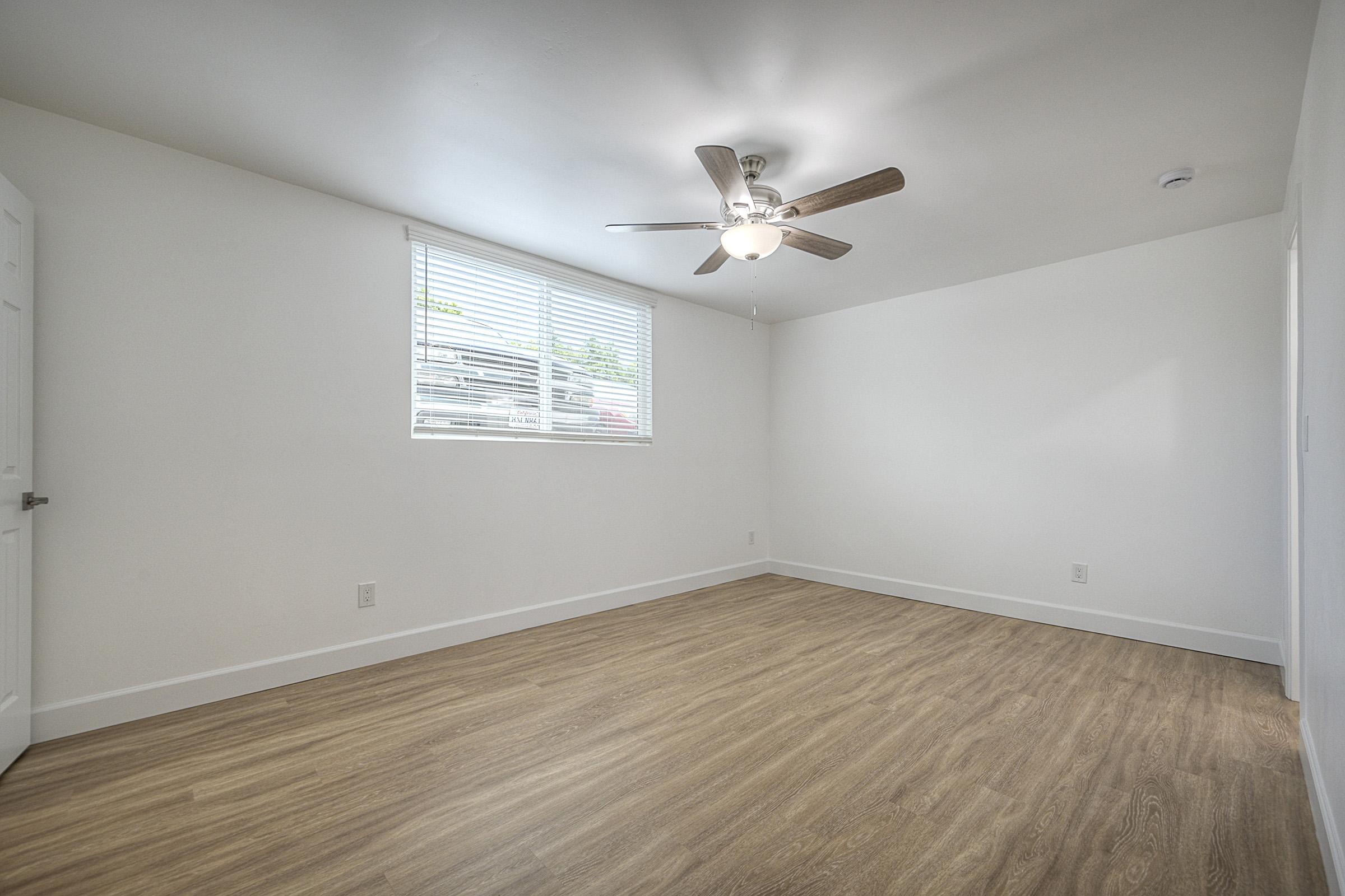 Empty room with light-colored walls and wooden flooring. A ceiling fan is installed, and there's a window with blinds allowing natural light to enter. The space appears clean and minimalist, suitable for various uses or personalization. No furniture is present.