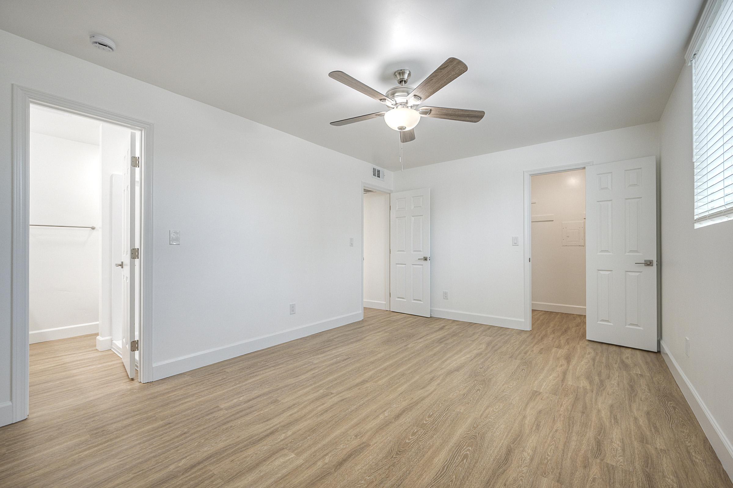 Bright and airy room with light-colored walls and a ceiling fan. Wood-like flooring adds warmth. Two doors lead to adjacent rooms, possibly closets or bathrooms. A window lets in natural light, enhancing the spacious feel of the area. Ideal for a living space or bedroom.