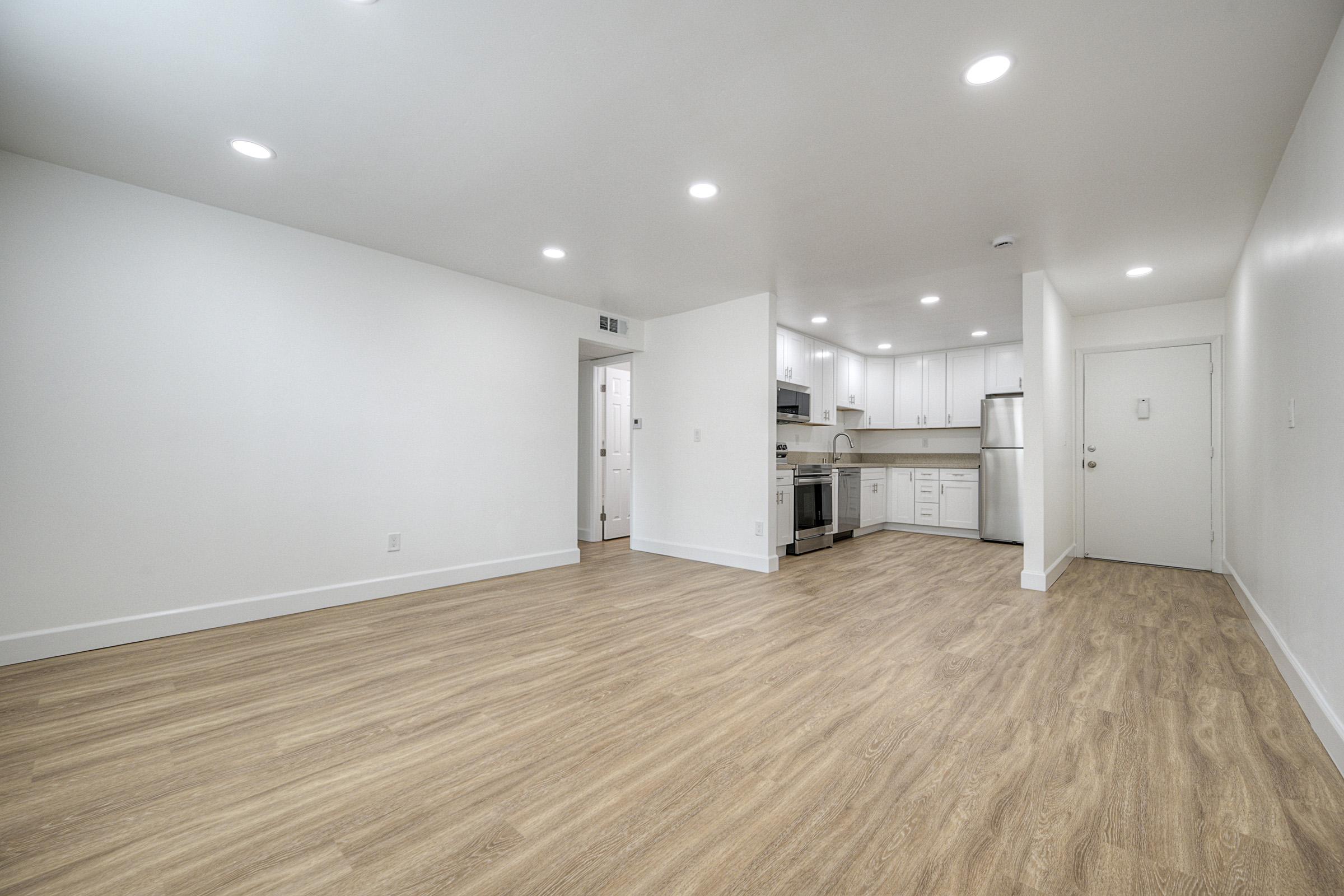 A spacious, modern kitchen and living area with light wood flooring, white walls, and recessed lighting. The kitchen features sleek appliances and cabinets. A doorway leads to a hallway while the entrance door is visible on the right side of the image.