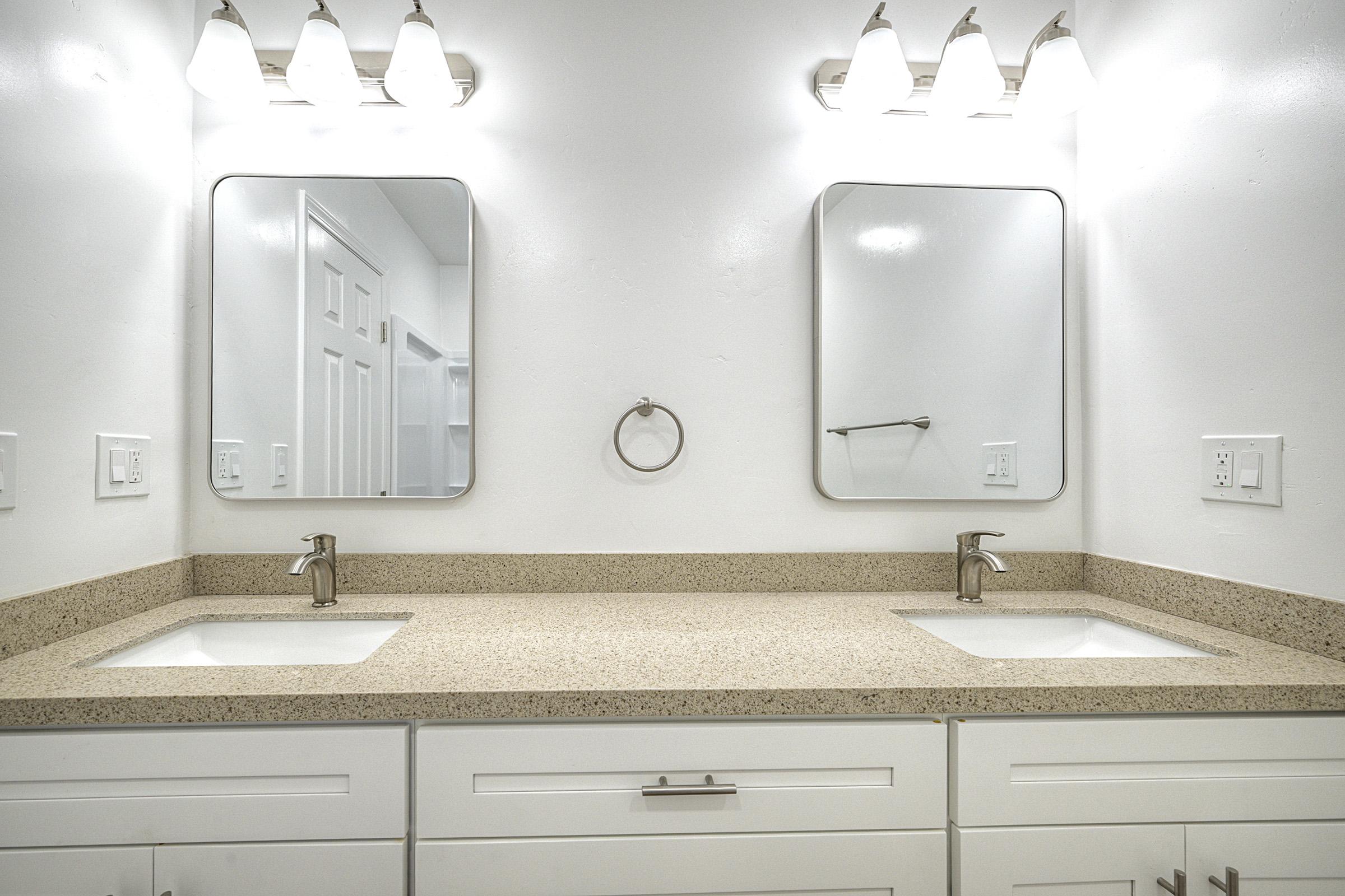 A modern bathroom featuring a double vanity with two sinks, each beneath a rectangular mirror. The countertop is a light speckled granite. Above the mirrors, there are four light fixtures providing bright illumination. The walls are painted white, and the space includes a towel ring and cabinet storage below the sinks.