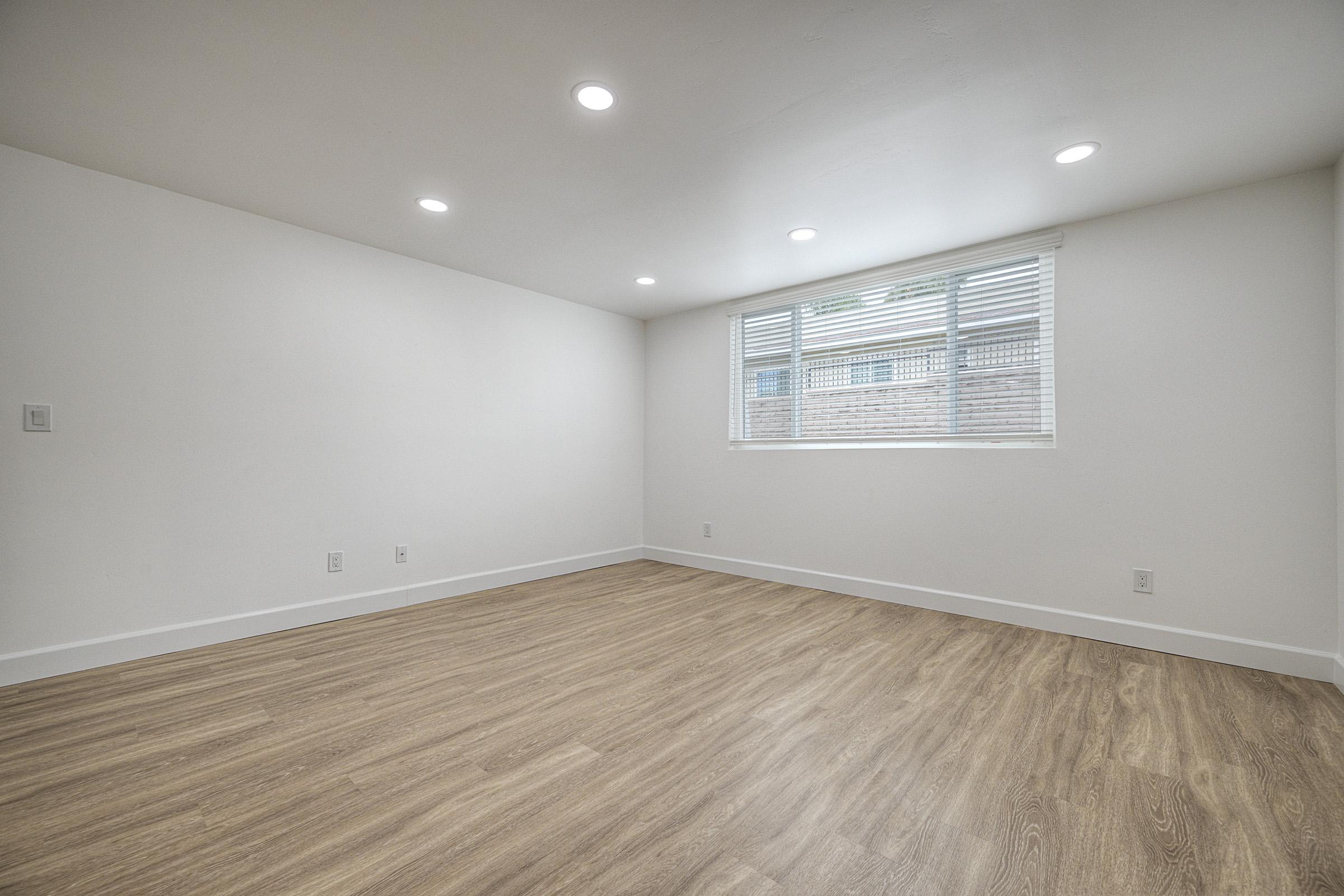 Empty room with light-colored walls and a wooden floor. The space features recessed lighting and a window with white blinds, allowing natural light to enter. There are no furnishings present, creating a clean and blank canvas for potential decoration or use.