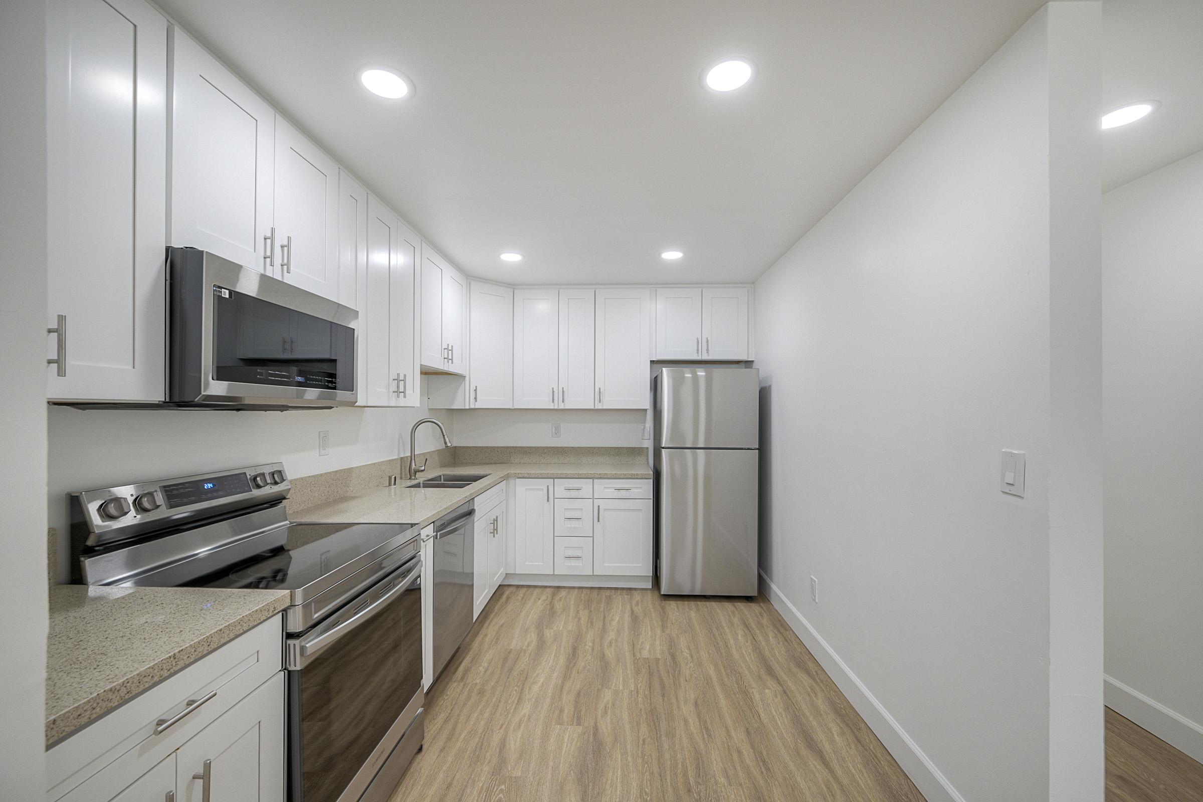 A modern kitchen featuring white cabinetry, stainless steel appliances including a microwave, oven, and refrigerator, along with a light-colored countertop. The space is bright, illuminated by recessed lighting, and has a warm wood floor. The design emphasizes a clean, contemporary aesthetic.