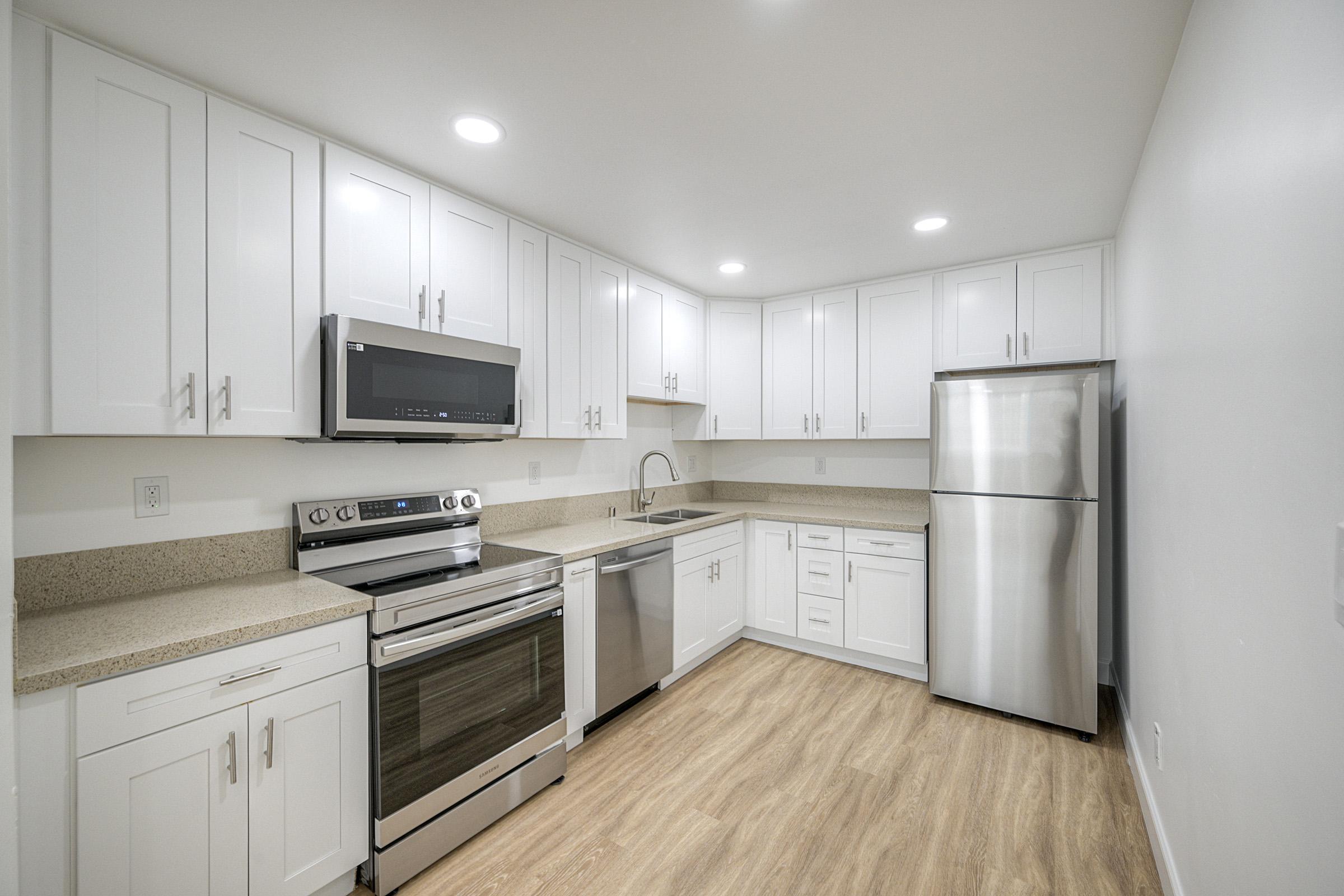 A modern kitchen featuring white cabinetry, stainless steel appliances, including a microwave, oven, and refrigerator. The countertops are light-colored with a textured finish, and the floor is a warm, light wood laminate. Bright lighting enhances the spacious and clean appearance of the kitchen.