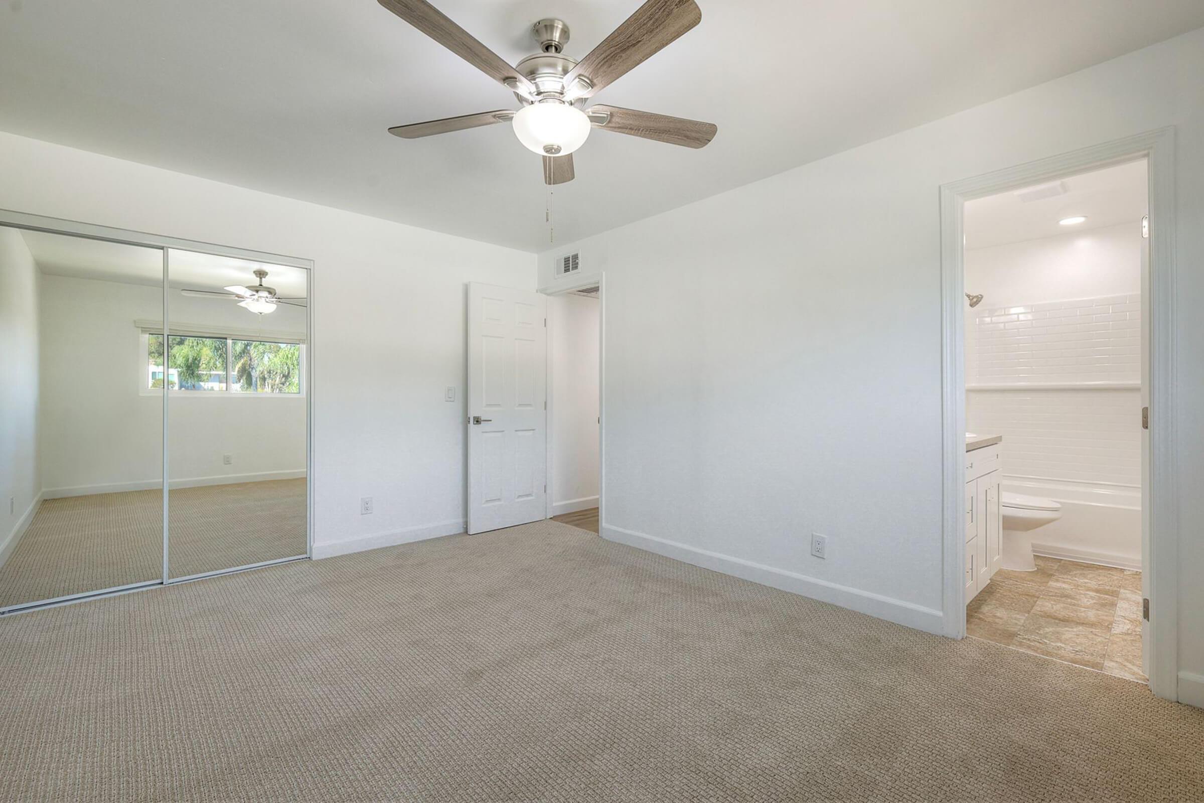 Spacious, well-lit bedroom featuring a ceiling fan, beige carpet, and mirrored closet doors. A doorway leads to a bathroom with white tiled walls, and a window allows natural light to fill the room. The walls are painted white, creating a fresh and airy atmosphere.