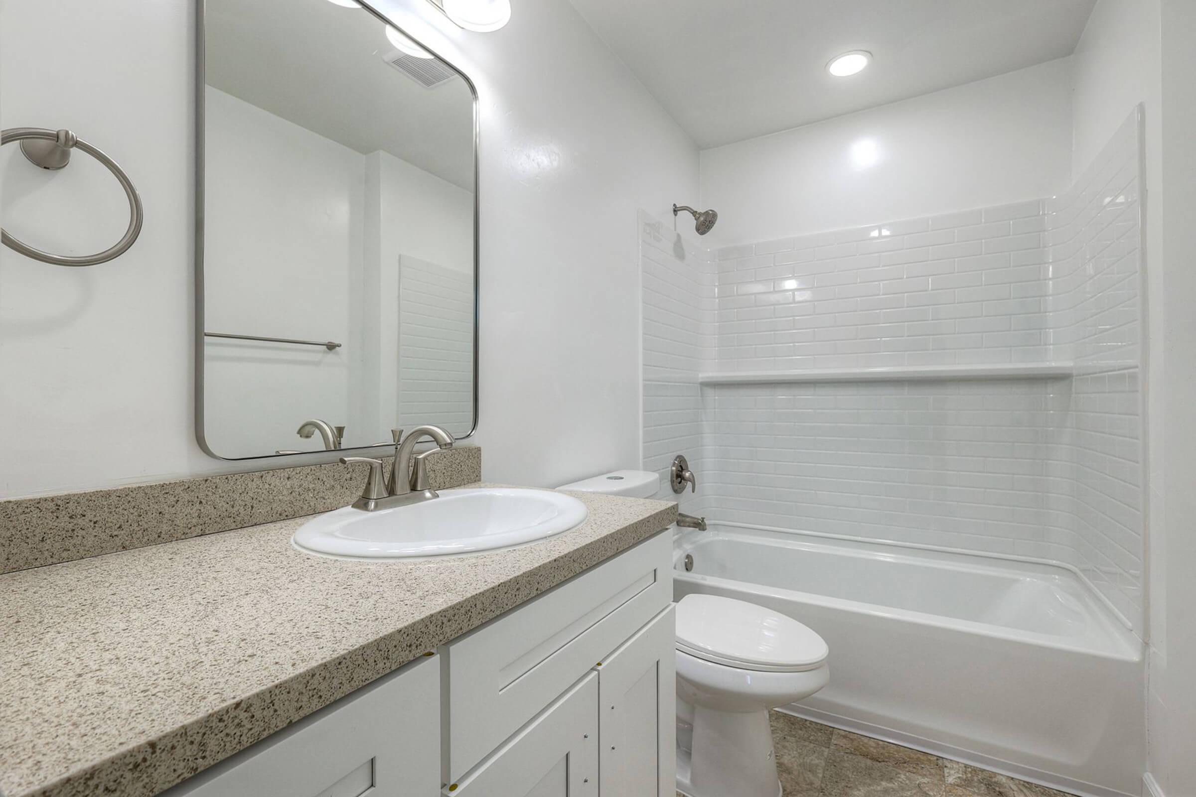 A modern bathroom featuring a clean, white tile shower and bathtub. It has a light-colored granite countertop with a sink, a mirror above the sink, and a towel bar on the wall. The walls are painted white, and there is bright overhead lighting, creating an airy and spacious feel.