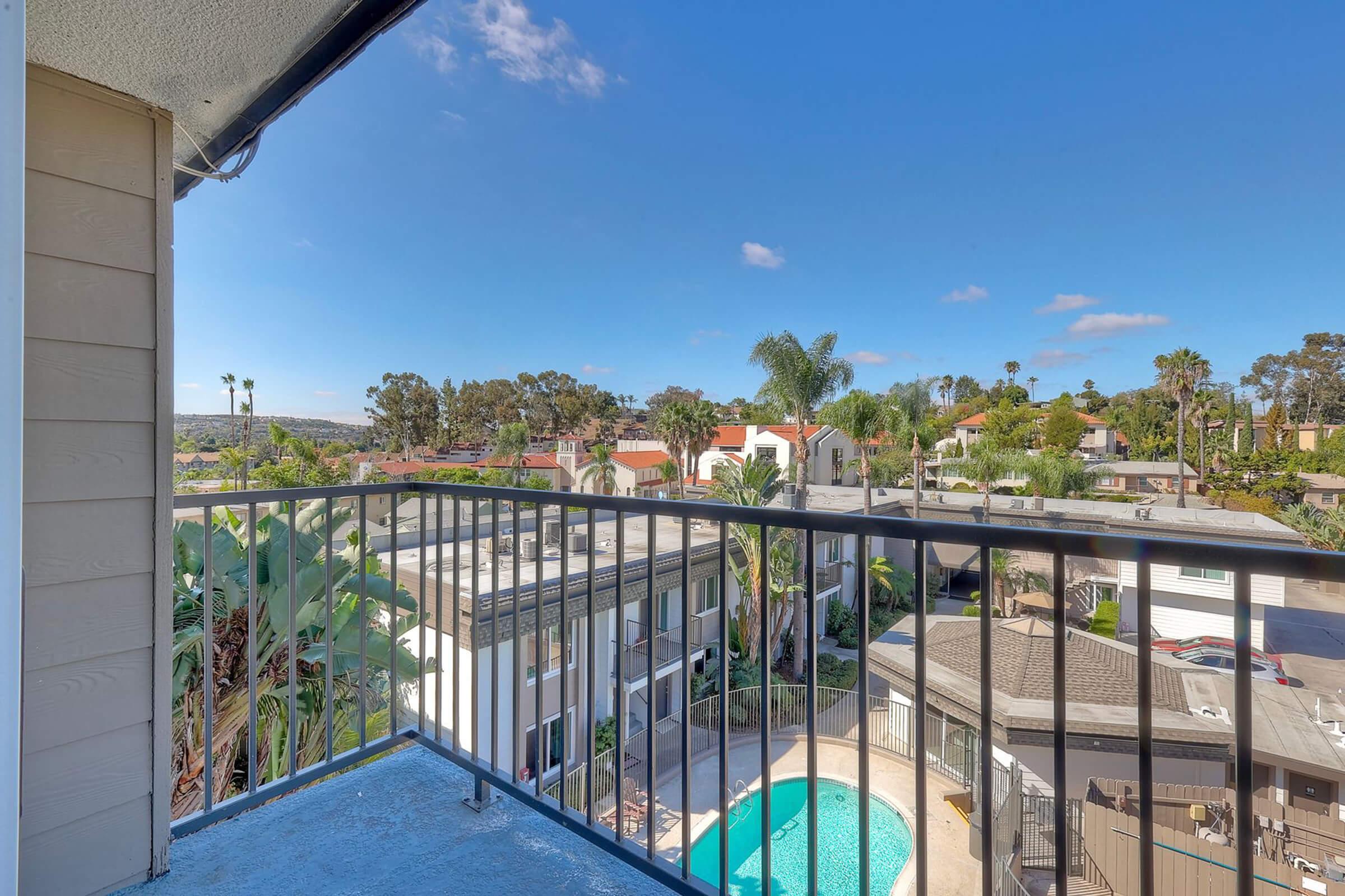 View from a balcony overlooking a pool and landscaped area, surrounded by palm trees and residential buildings under a bright blue sky.