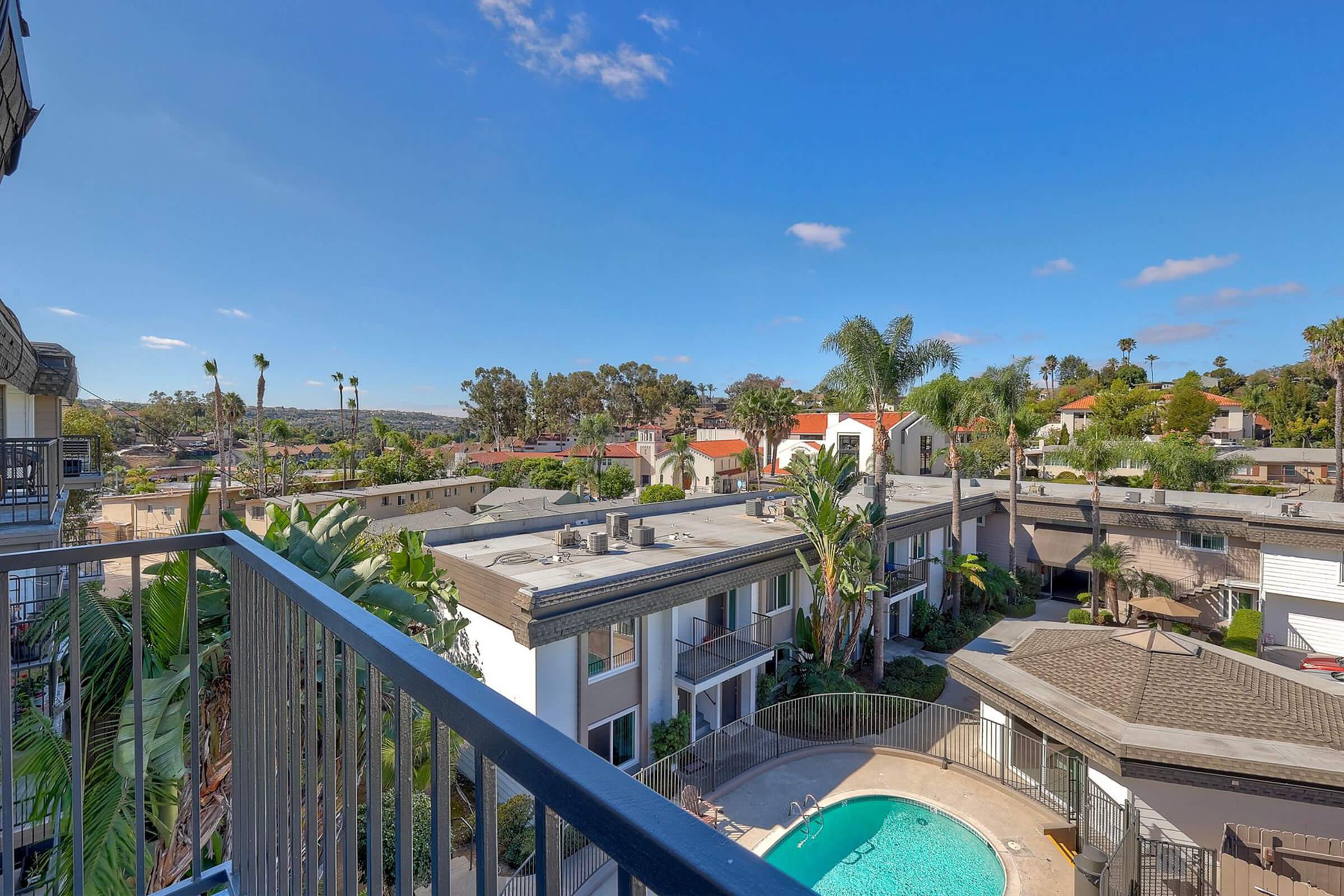 View from a balcony overlooking a tropical landscape with palm trees and a swimming pool. The scene includes various buildings, a clear blue sky, and residential rooftops in the background, creating a vibrant, sunny atmosphere.
