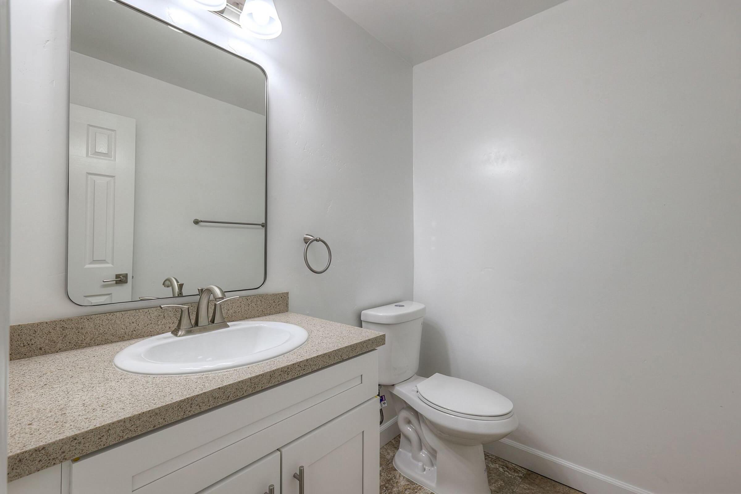 A clean, modern bathroom featuring a white vanity with a sink, a large mirror above the sink, a toilet, and light-colored walls. The floor has a neutral tile pattern, and there is a towel ring mounted on the wall. The overall atmosphere is bright and minimalistic.