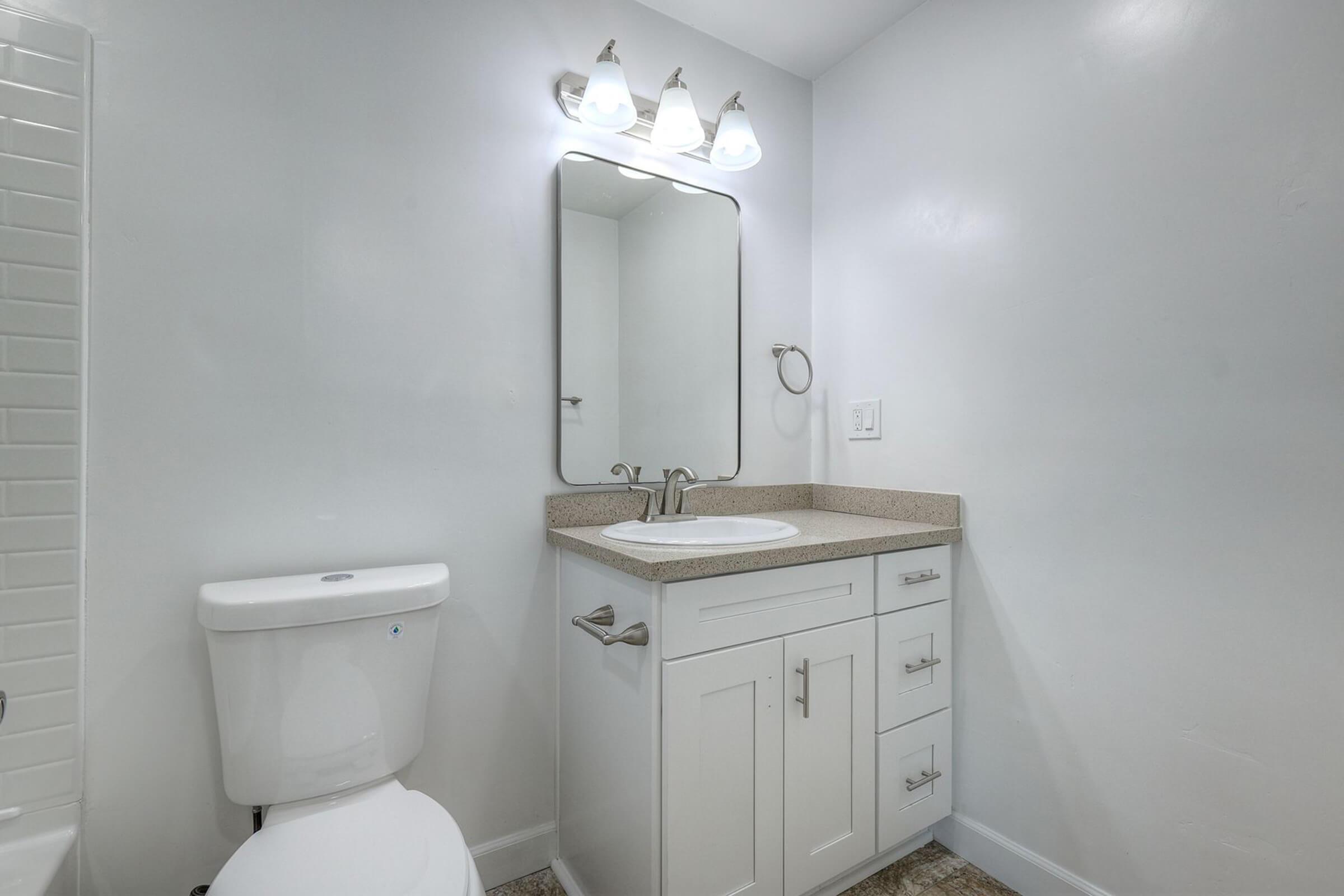 A clean and modern bathroom featuring a white toilet, a single-sink vanity with a countertop, and a large mirror. The walls are painted white, and three light fixtures are mounted above the mirror, providing bright illumination. The overall look is sleek and contemporary.