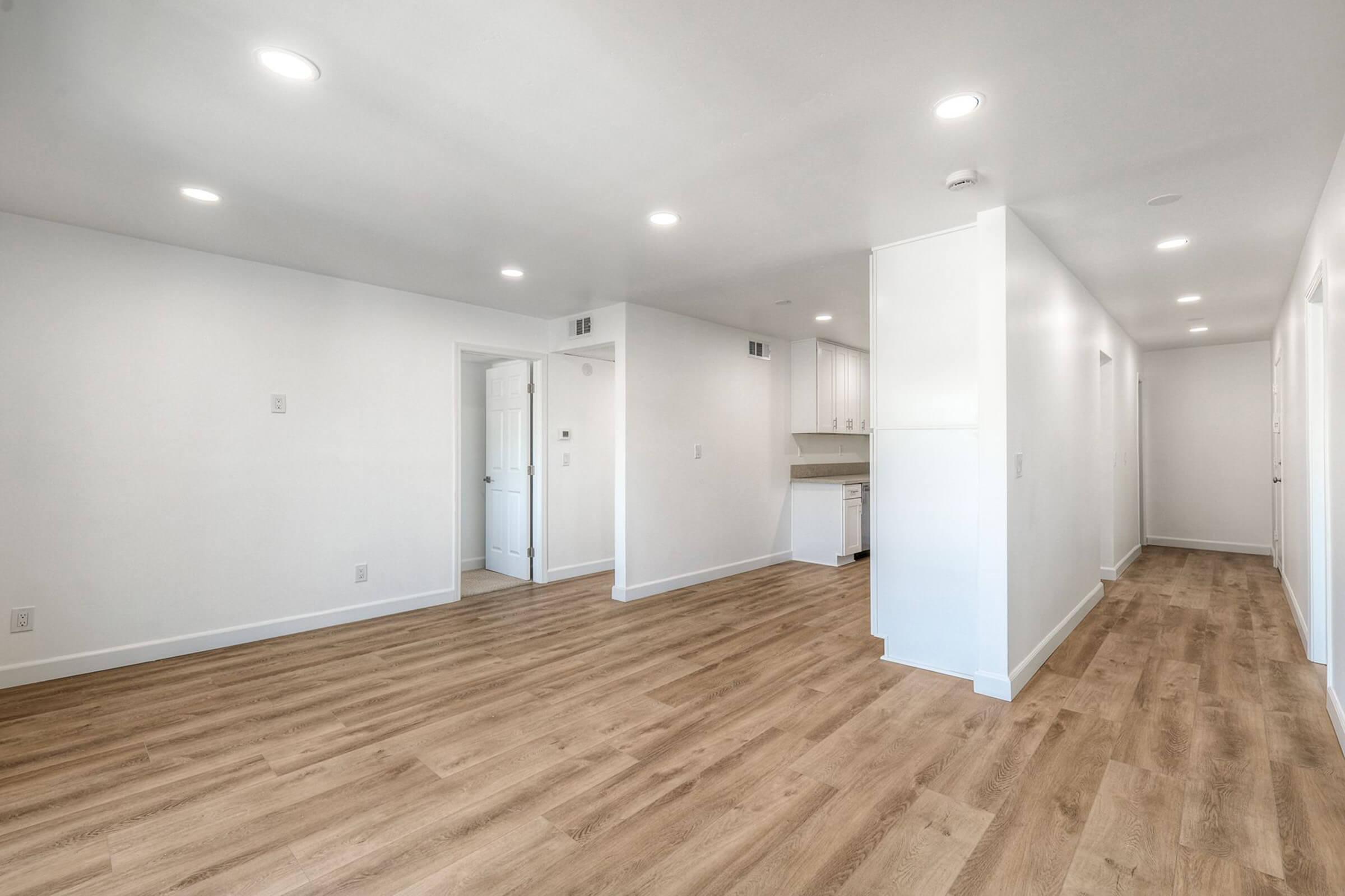 A modern, open-concept interior space featuring light-colored walls and a hardwood floor. The area is well-lit with recessed ceiling lights. On the left, there is a kitchen with white cabinetry, while empty doorways lead to other rooms along the right side.