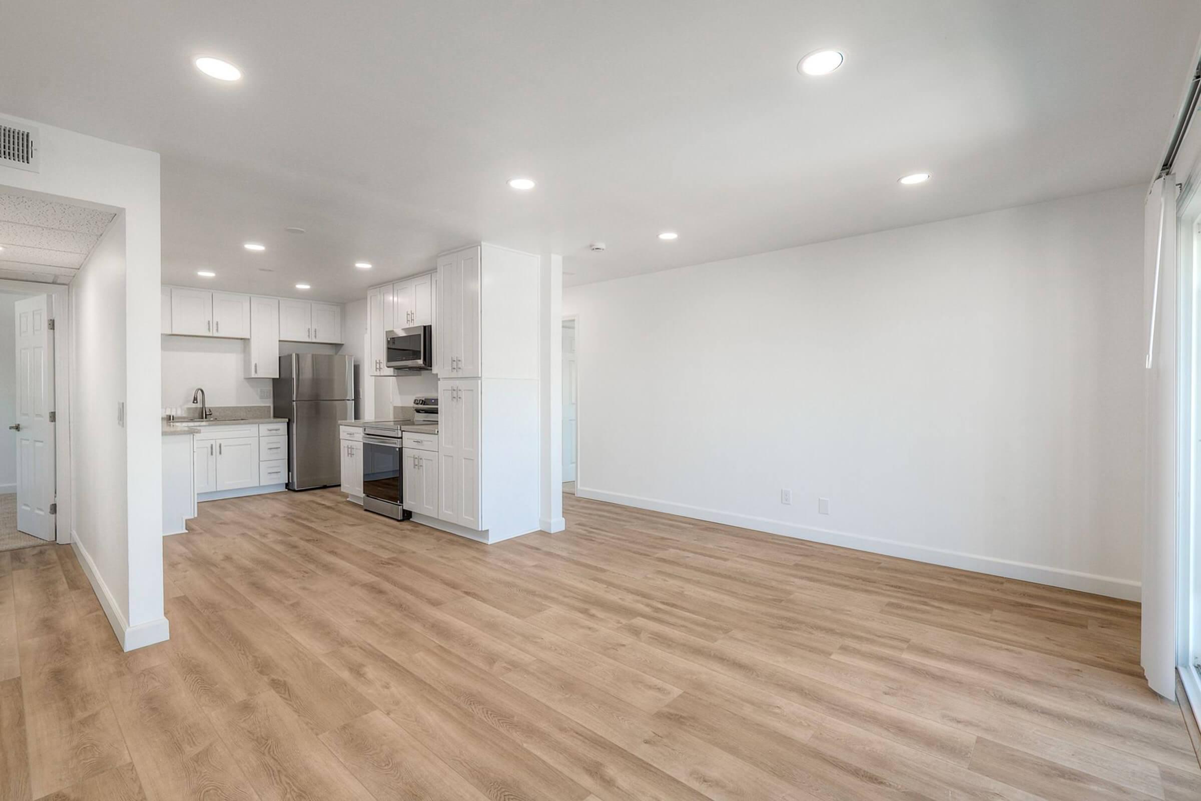 A modern, empty kitchen and living area featuring light wooden flooring, white walls, and ample natural light. The kitchen has stainless steel appliances, white cabinets, and a countertop, while the living space is spacious and uncluttered, with sliding doors leading to an outside area.