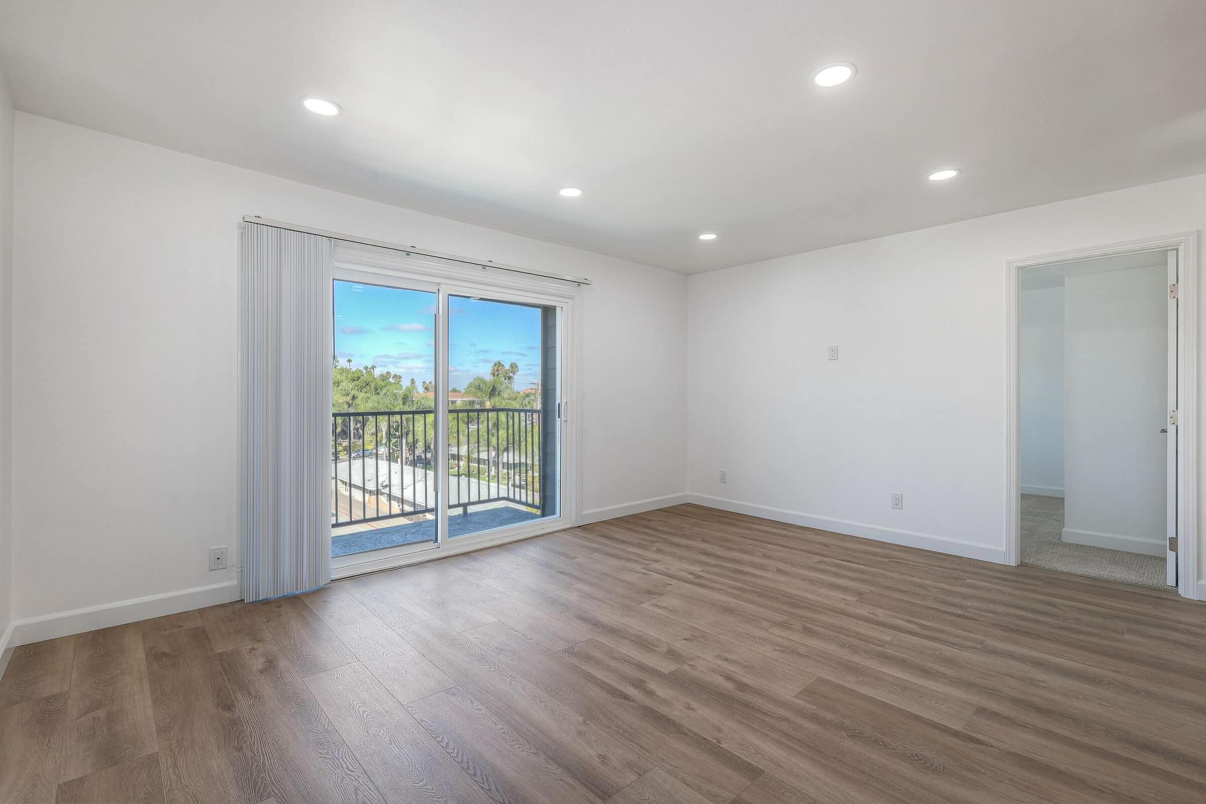 A spacious, empty room with light wooden flooring and white walls. There are large sliding glass doors leading to a balcony, allowing natural light to fill the space. The ceiling has recessed lighting, and there is a doorway to another room on the right. The atmosphere is bright and airy, ideal for interior design possibilities.