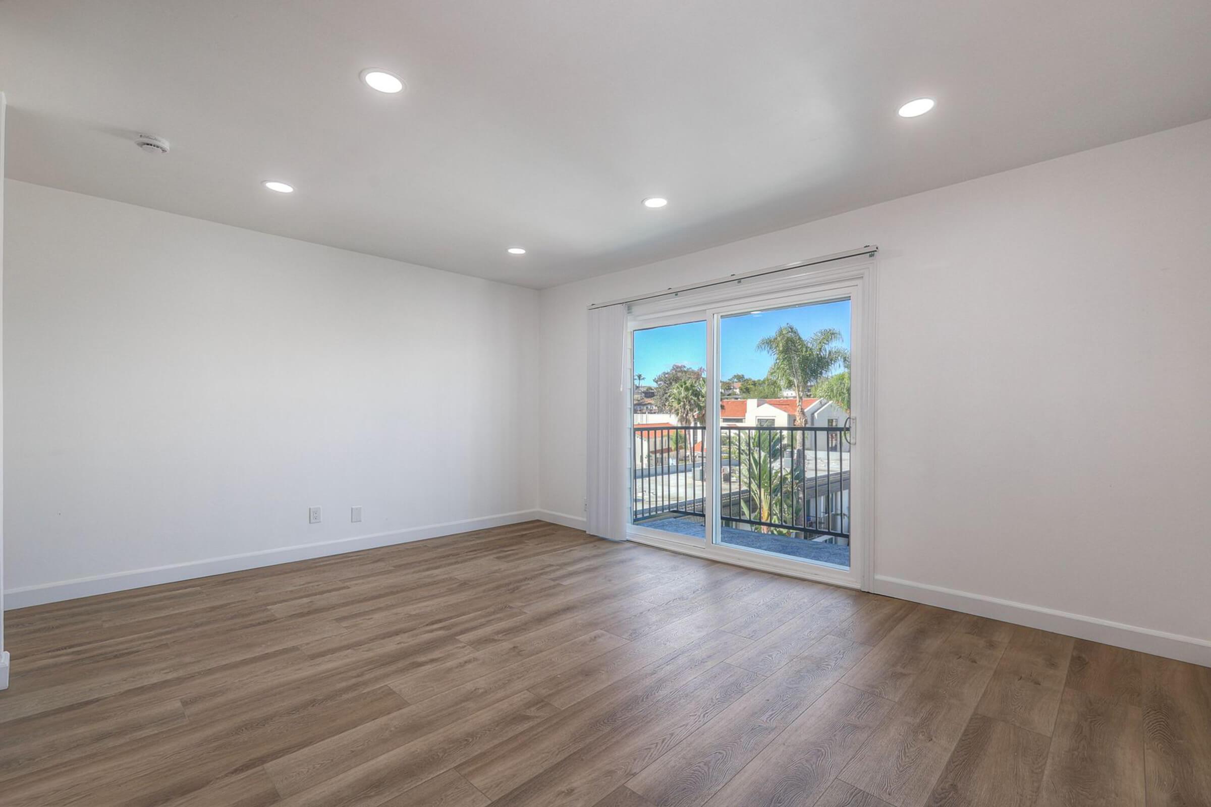 A spacious, empty room with light-colored walls and wooden flooring. There are large sliding glass doors leading to a balcony with a view of palm trees outside. The ceiling is equipped with recessed lighting, creating a bright and airy atmosphere.