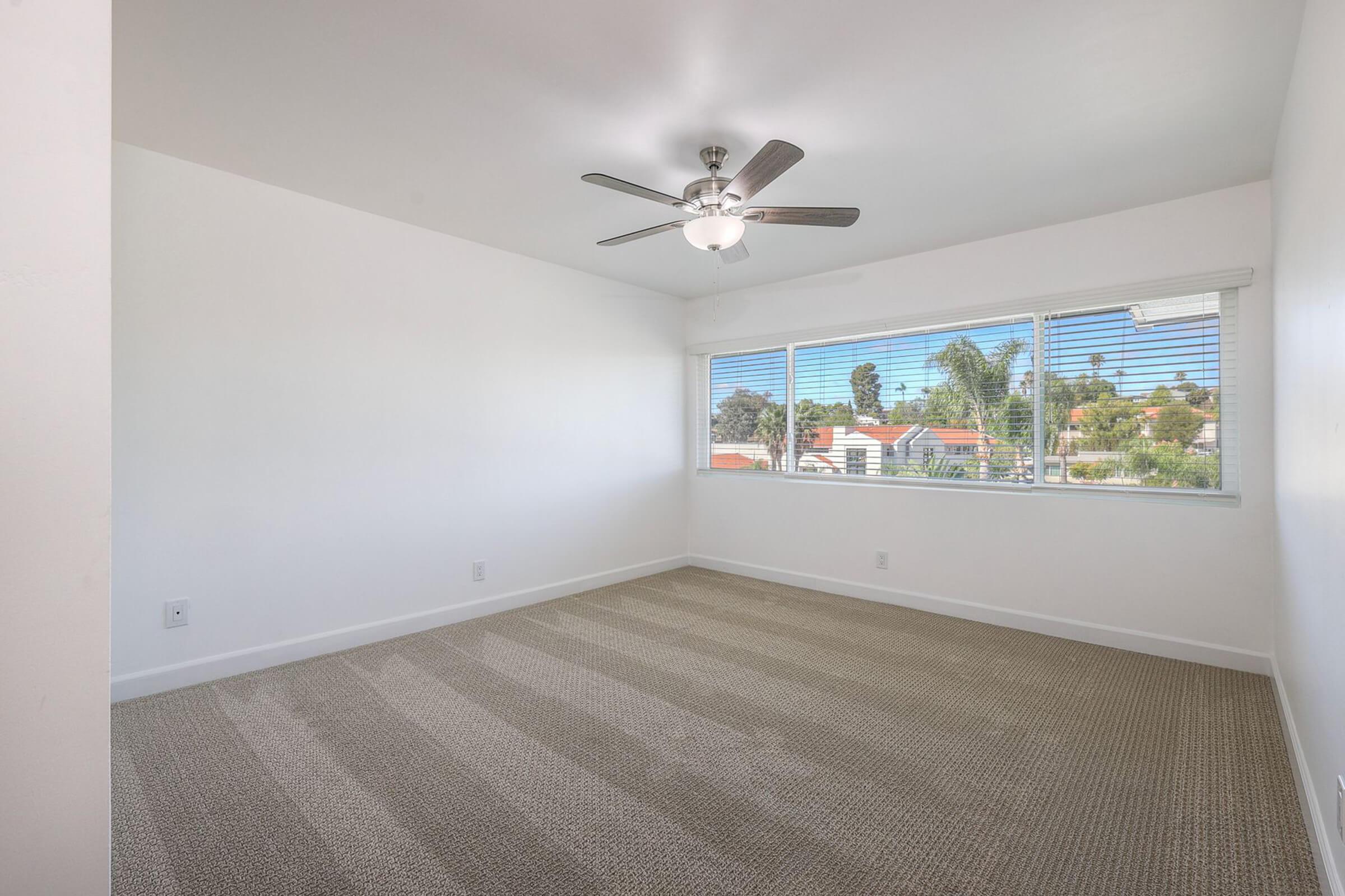 A bright, empty room featuring light-colored walls and a ceiling fan. Large windows allow for natural light and offer a view of greenery outside. The floor is covered in a light beige carpet with subtle stripes, creating a clean and spacious atmosphere.
