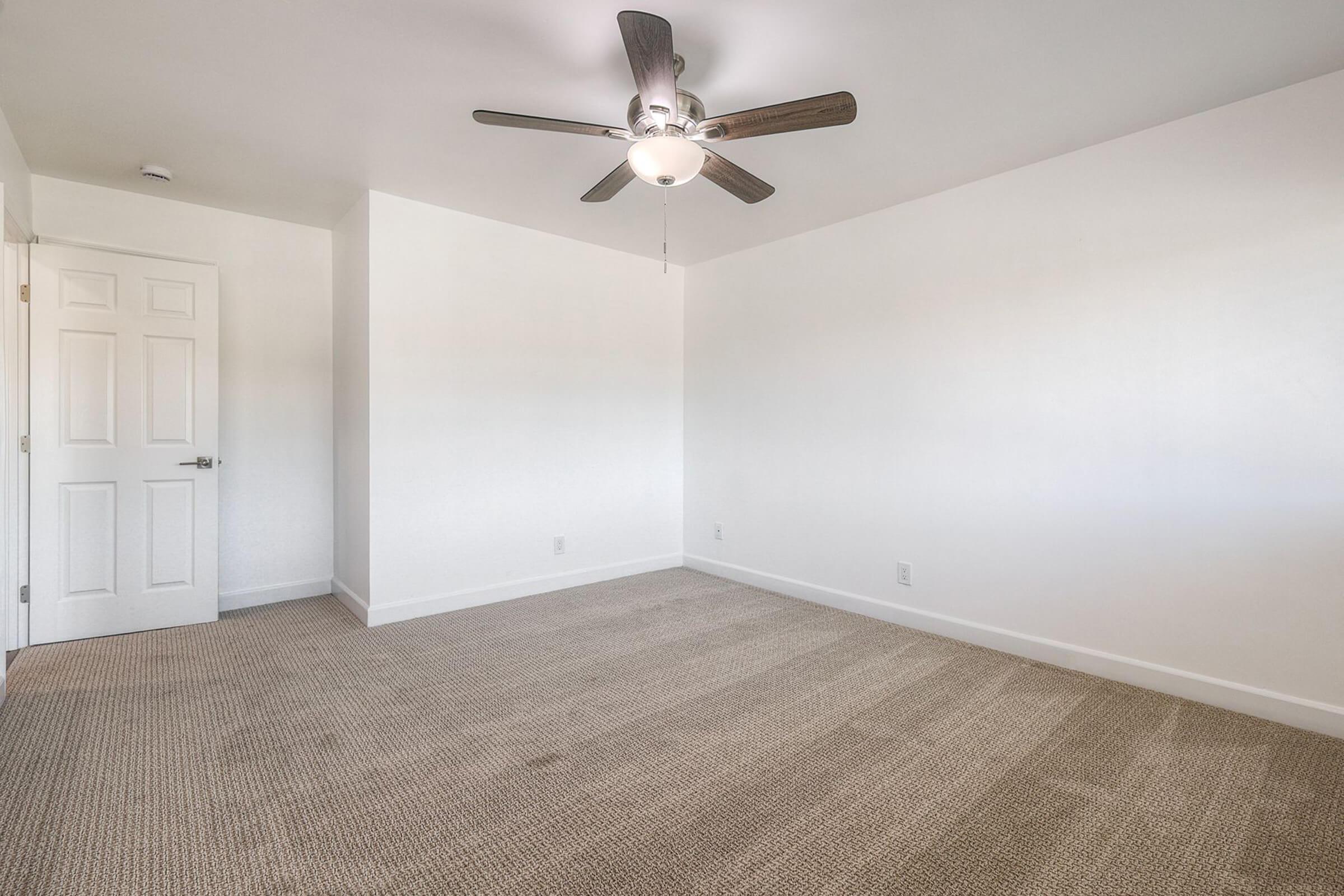 A clean, empty room with light beige carpet and white walls. A ceiling fan with wooden blades is mounted in the center, and there’s a closed white door on the left side of the image. The room is well-lit and appears spacious, with natural light coming from an unseen source.