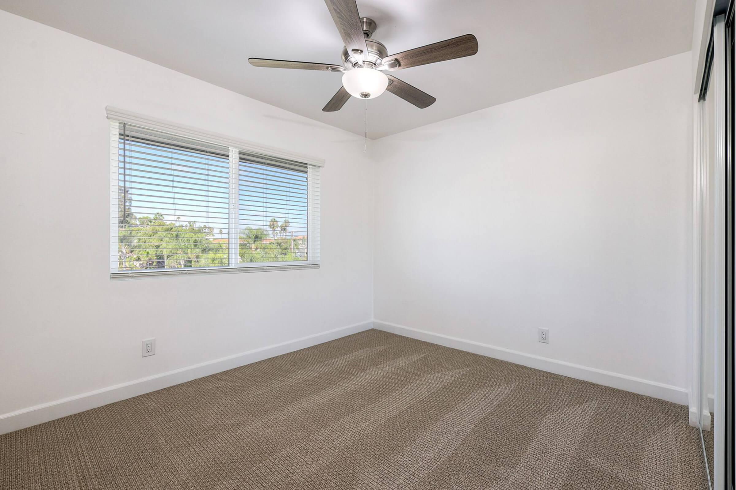 A bright, empty bedroom featuring a ceiling fan and large window with blinds. The walls are painted white, and the floor is covered with beige carpet. There are no furnishings, creating a minimalist and spacious atmosphere.