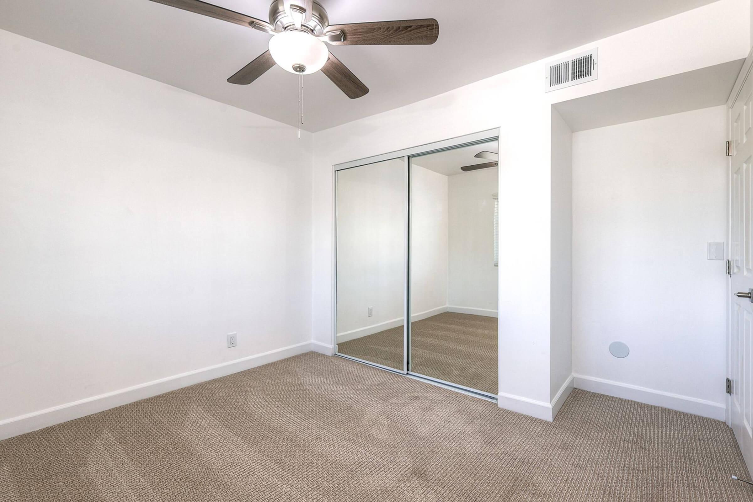 A small, empty room with light-colored walls and carpeted flooring. It features a ceiling fan with dark blades, a mirrored closet with sliding doors, and a small wall-mounted speaker. Natural light streams in through a window, adding brightness to the space.